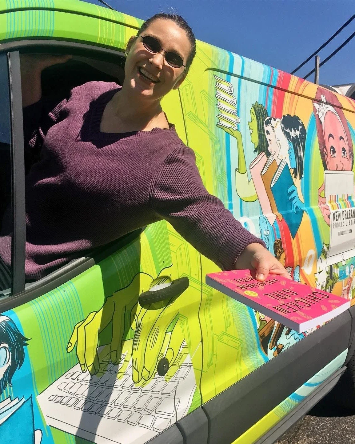 A smiling woman with sunglasses in a purple top holding a pink book, leaning out of a colorful vehicle with cartoon illustrations of people and a keyboard image on the side, during daytime.