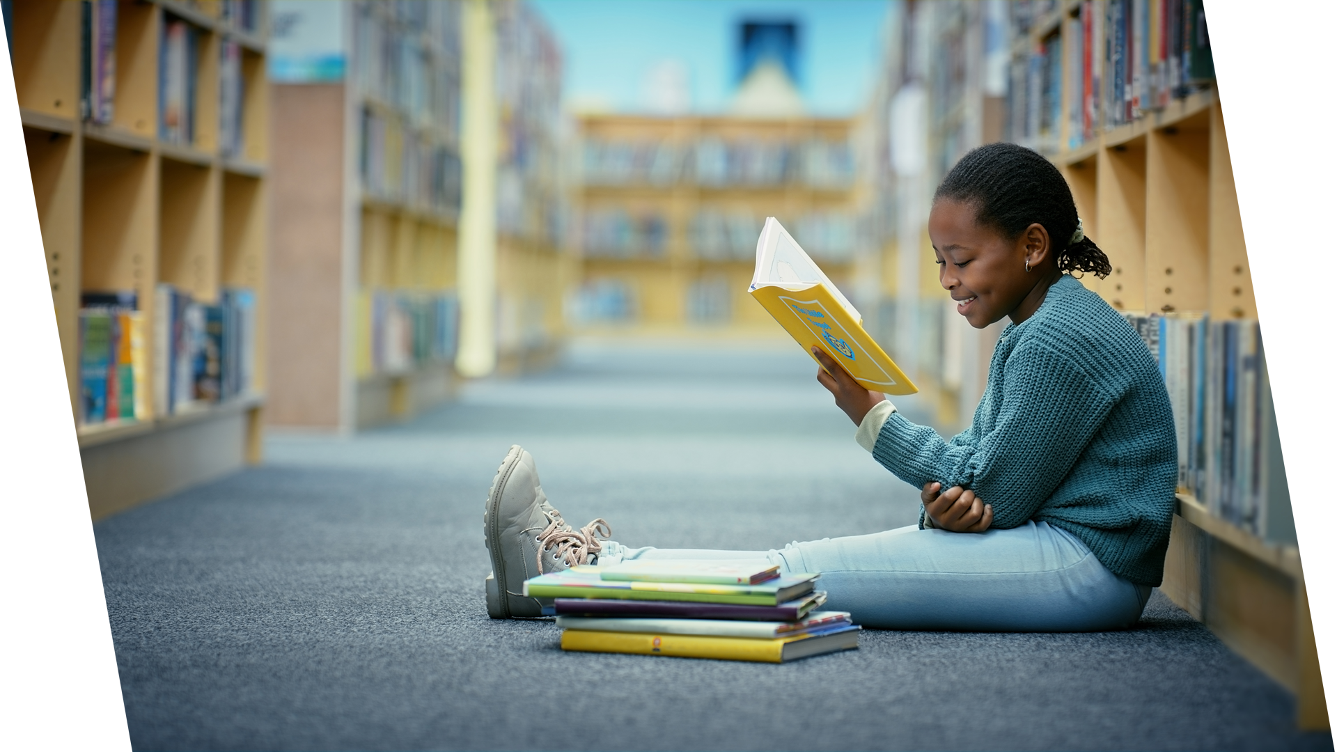 A young girl sitting on the floor in a library, reading a yellow book, with a stack of books beside her.