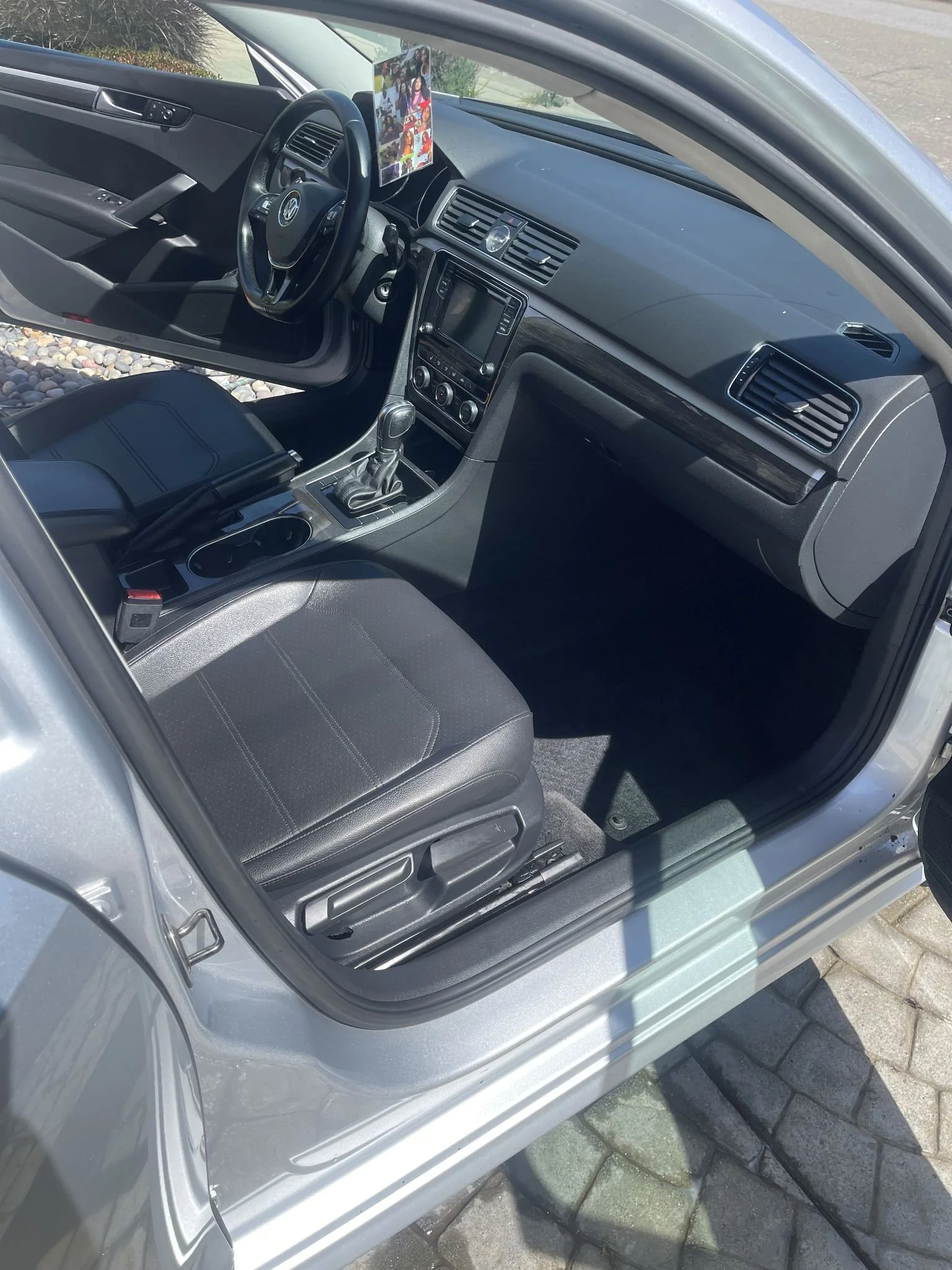 Interior of a silver Volkswagen car showing the driver's seat, dashboard, steering wheel, gear shift, and a mounted phone holder with photos displayed.