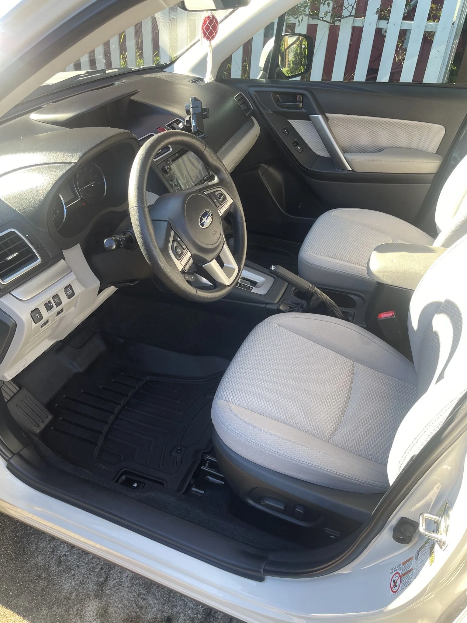 Interior of a white car showing the front seats, steering wheel, dashboard, and part of the door panel. The seats are light gray with a textured fabric, and the dashboard is black with various controls.