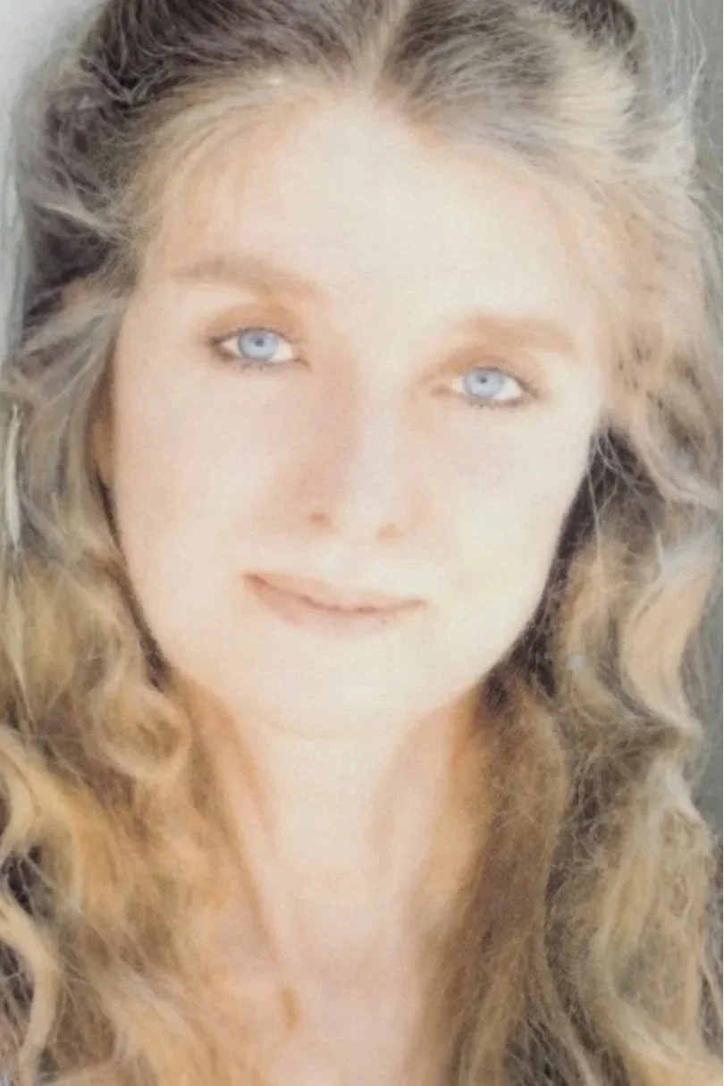 Close-up of a woman with long, curly blonde hair and light blue eyes, smiling softly.