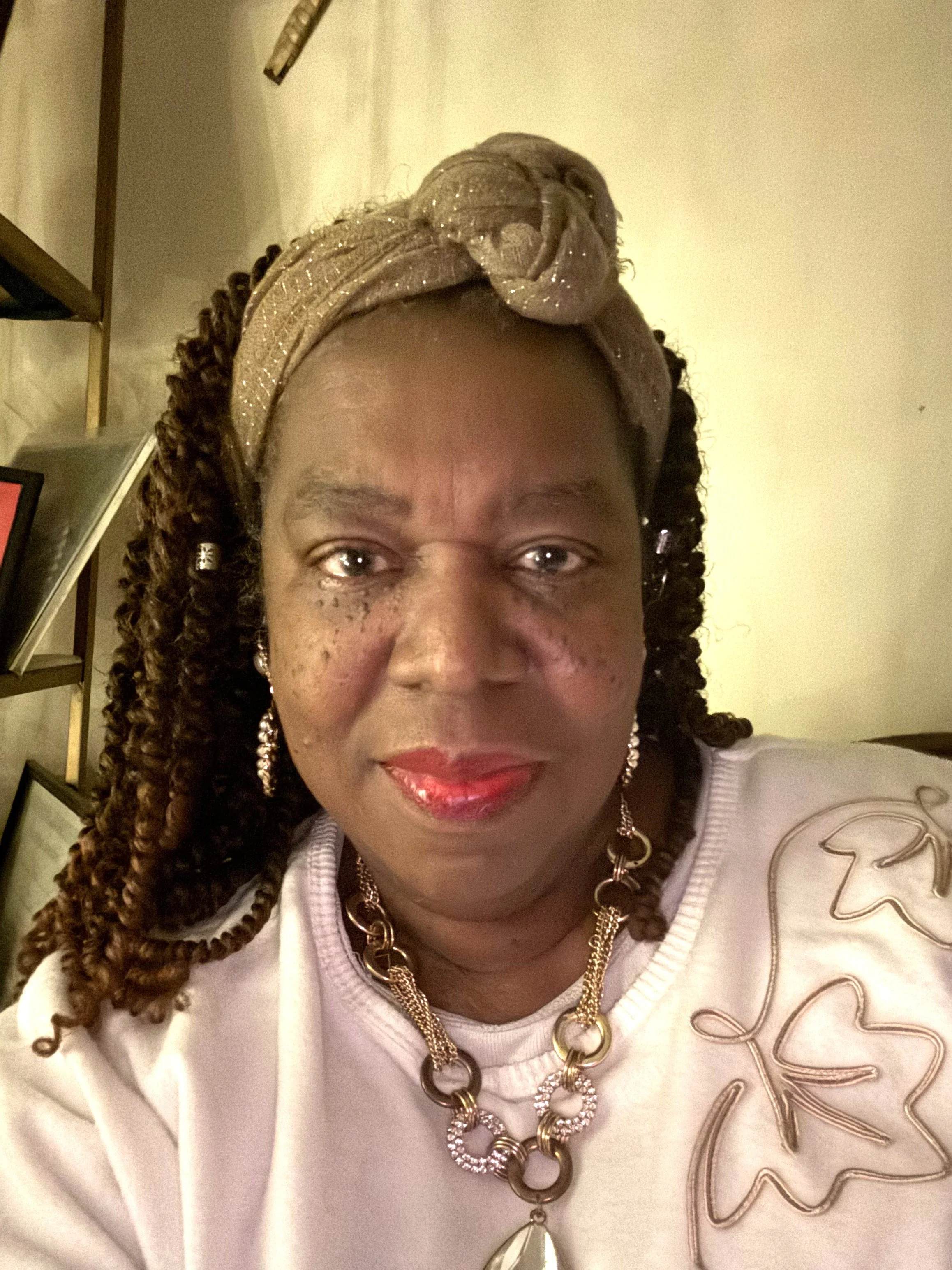 A woman with dark curly hair wearing a beige headwrap, gold jewelry, and a cream-colored shirt with gold embroidery.