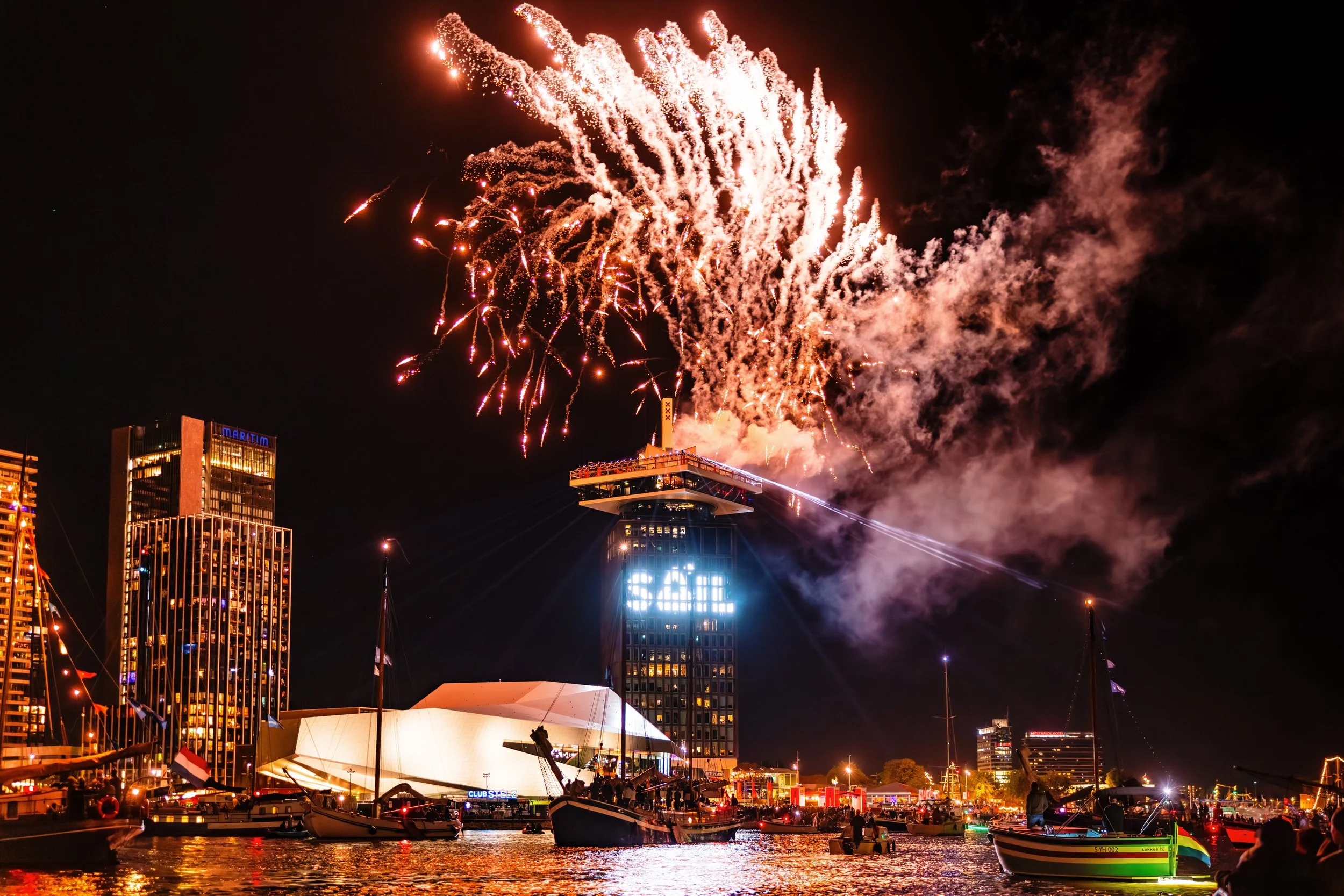 Vuurwerkshow tijdens de dagafsluiting van SAIL 2025 vanaf het dak van de ADAM toren, vuurwerk explodeert boven een haven met boten en hoge gebouwen in de stad 's nachts.