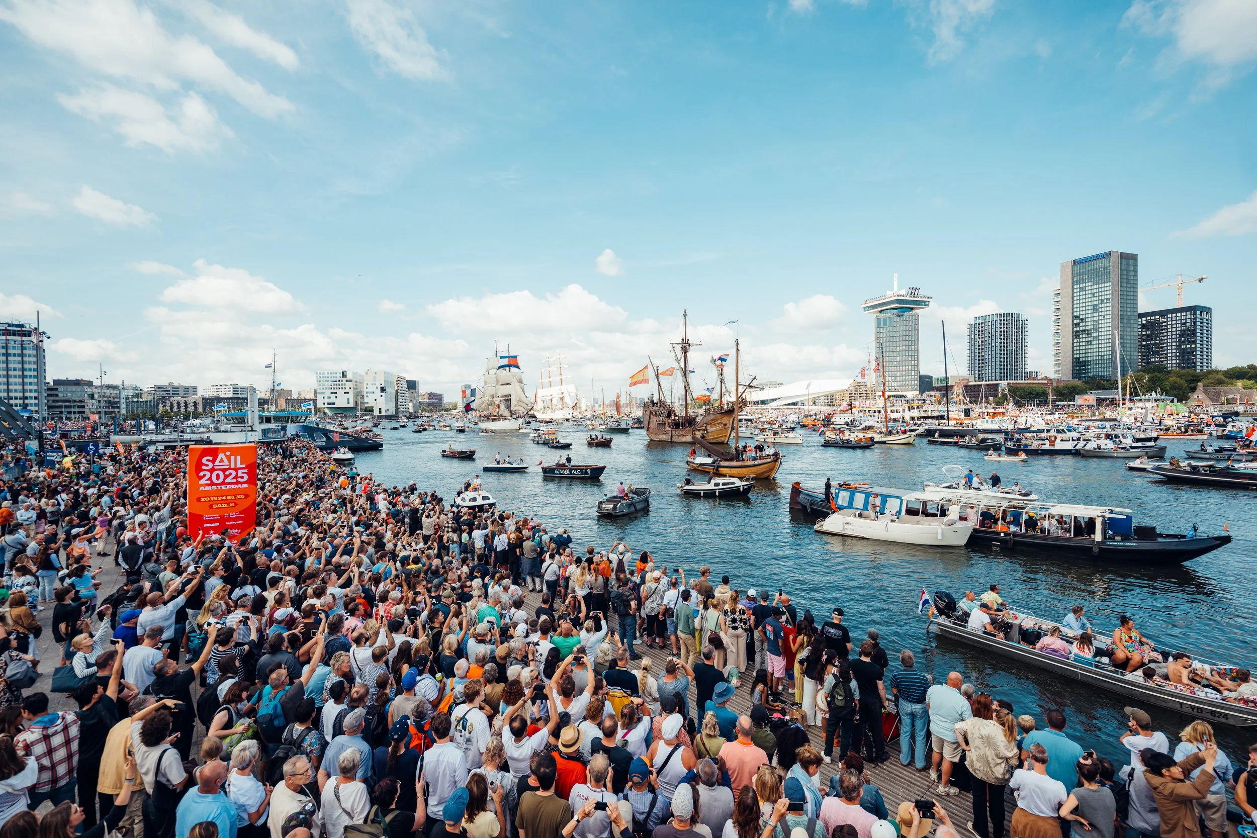 Een drukke menigte mensen tijdens SAIL 2025 langs de kade van een haven met verschillende historische schepen en moderne hoge gebouwen op de achtergrond, under een heldere blauw hemel met enkele wolken.