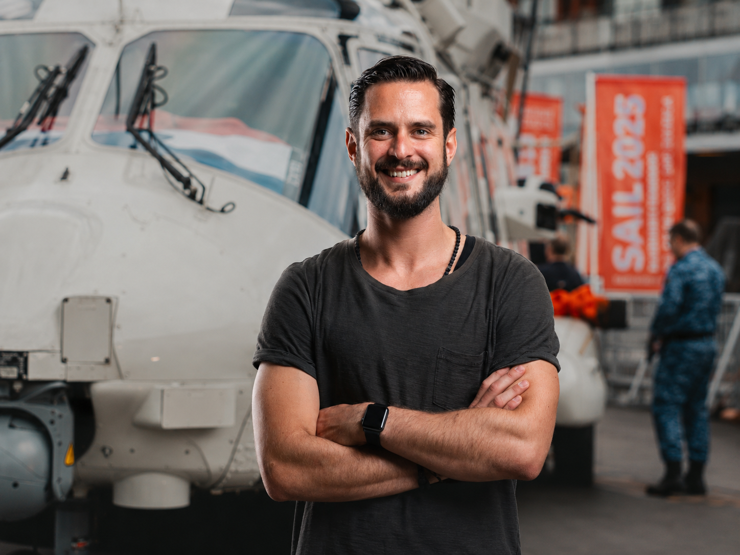 Een lachende man met een korte baard en zwart haar, gekleed in een zwart T-shirt, staat met gekruiste armen voor een helikopter in een vliegtuigloods.
