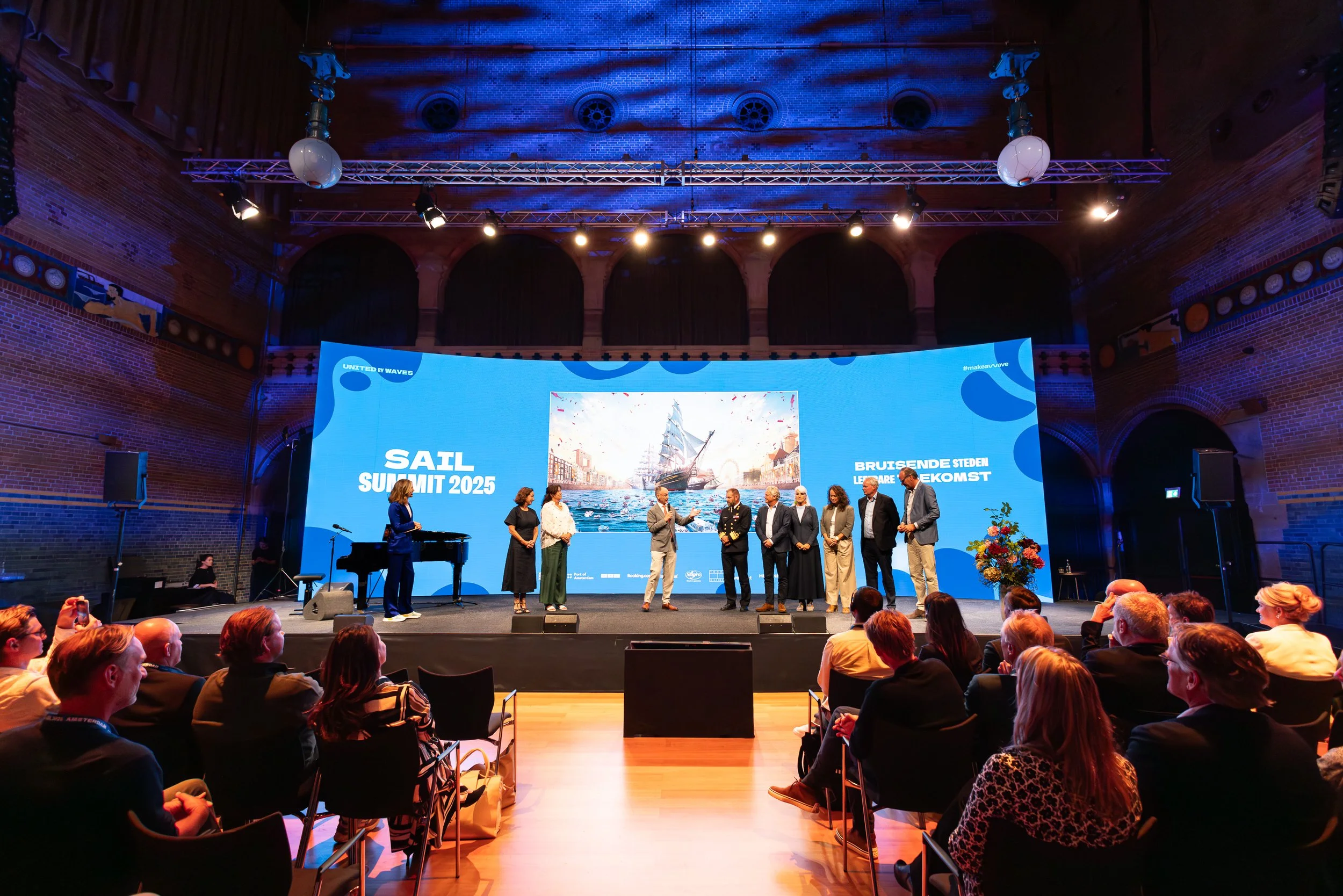 Een groep mensen staat op een podium tijdens de SAIL Summit 2025 in de Beurs van Berlage, met een groot scherm op de achtergrond dat een schip op de wateren toont, in een zaal vol mensen.