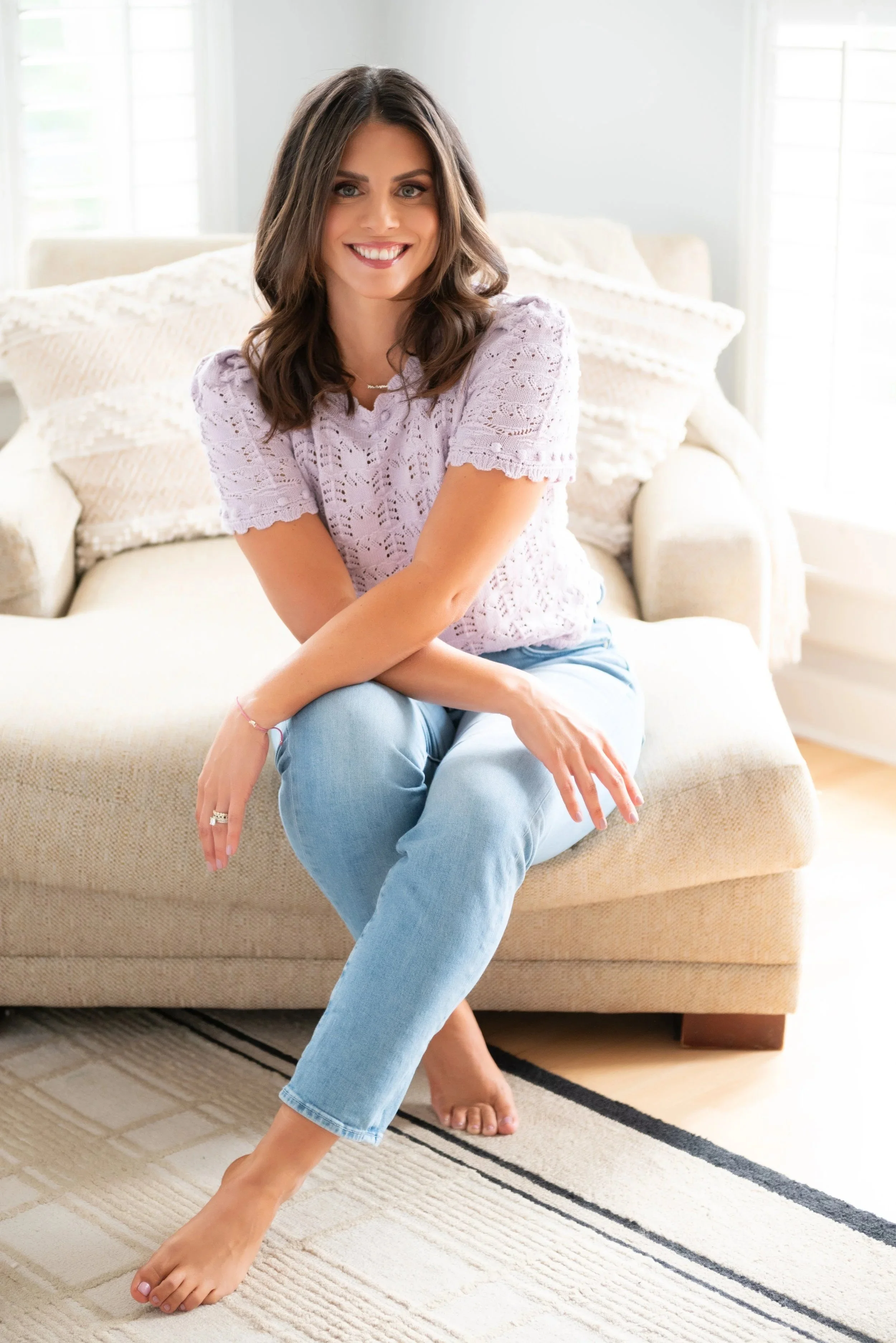 A woman with brown hair and a bright smile sitting on a beige couch in a well-lit room, wearing a lavender top and light blue jeans.