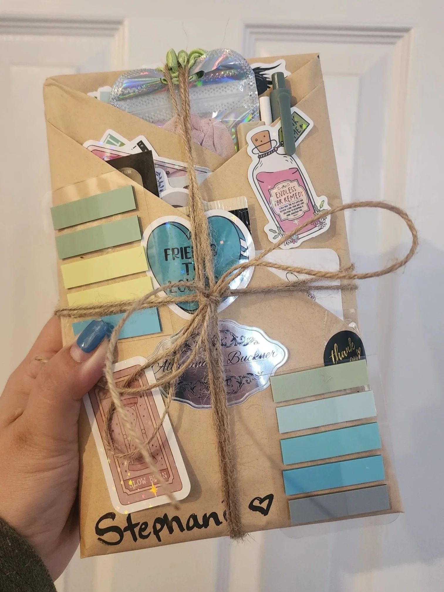 A hand holding a gift wrapped in brown paper and tied with twine, decorated with stickers, notes, and cards.