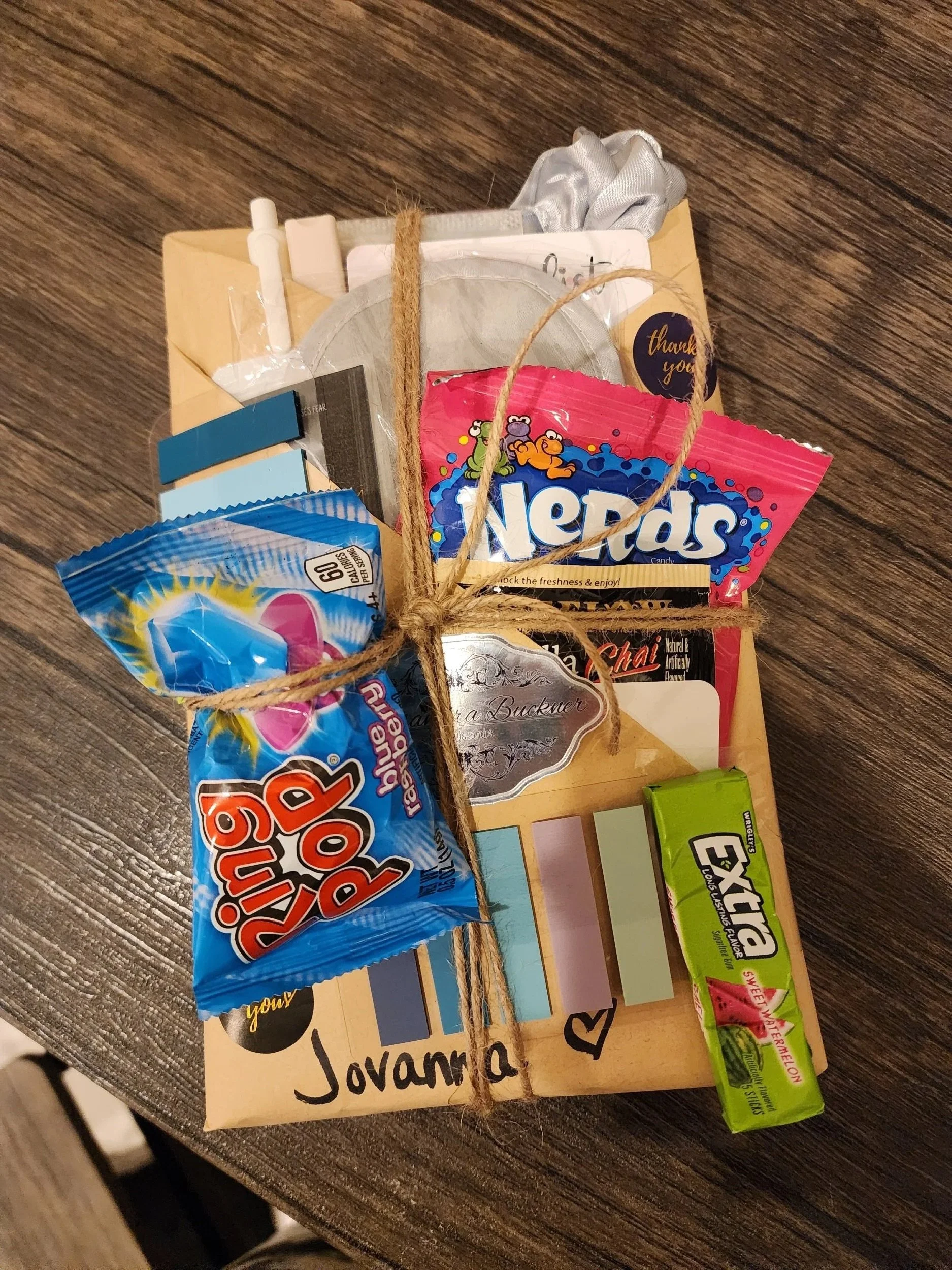 A gift box wrapped with a twine bow, containing various candies and snacks, including Nerds, Airheads, and Exta Juicy Watermelon gum, placed on a wooden surface with personalized writing and decorations on the box.
