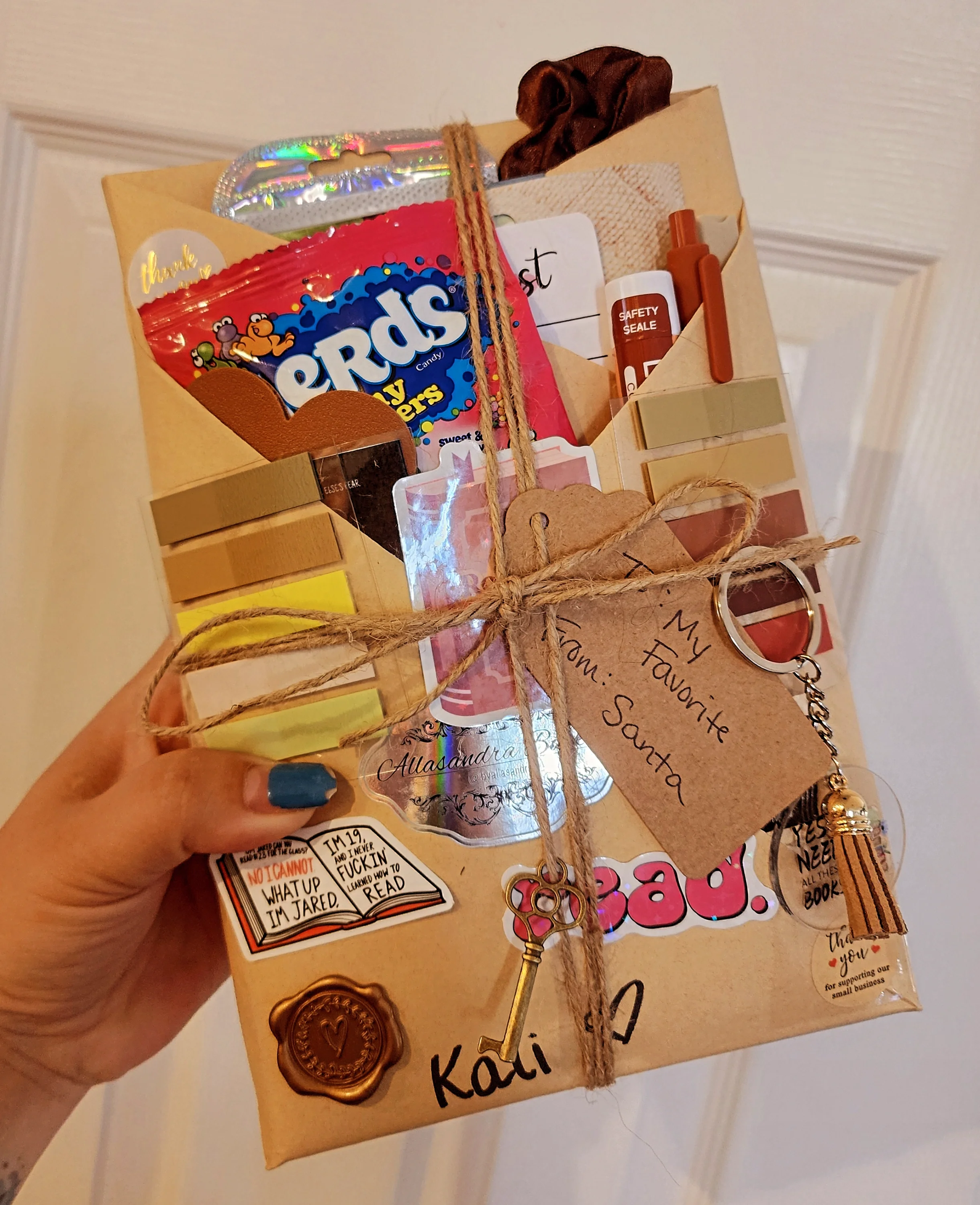 A gift box wrapped in brown paper and tied with twine, decorated with various stickers, a key, and a handwritten tag that reads 'My Favorite Mom: Santa.' The box contains snacks, stickers, a safety seal, and small notes.