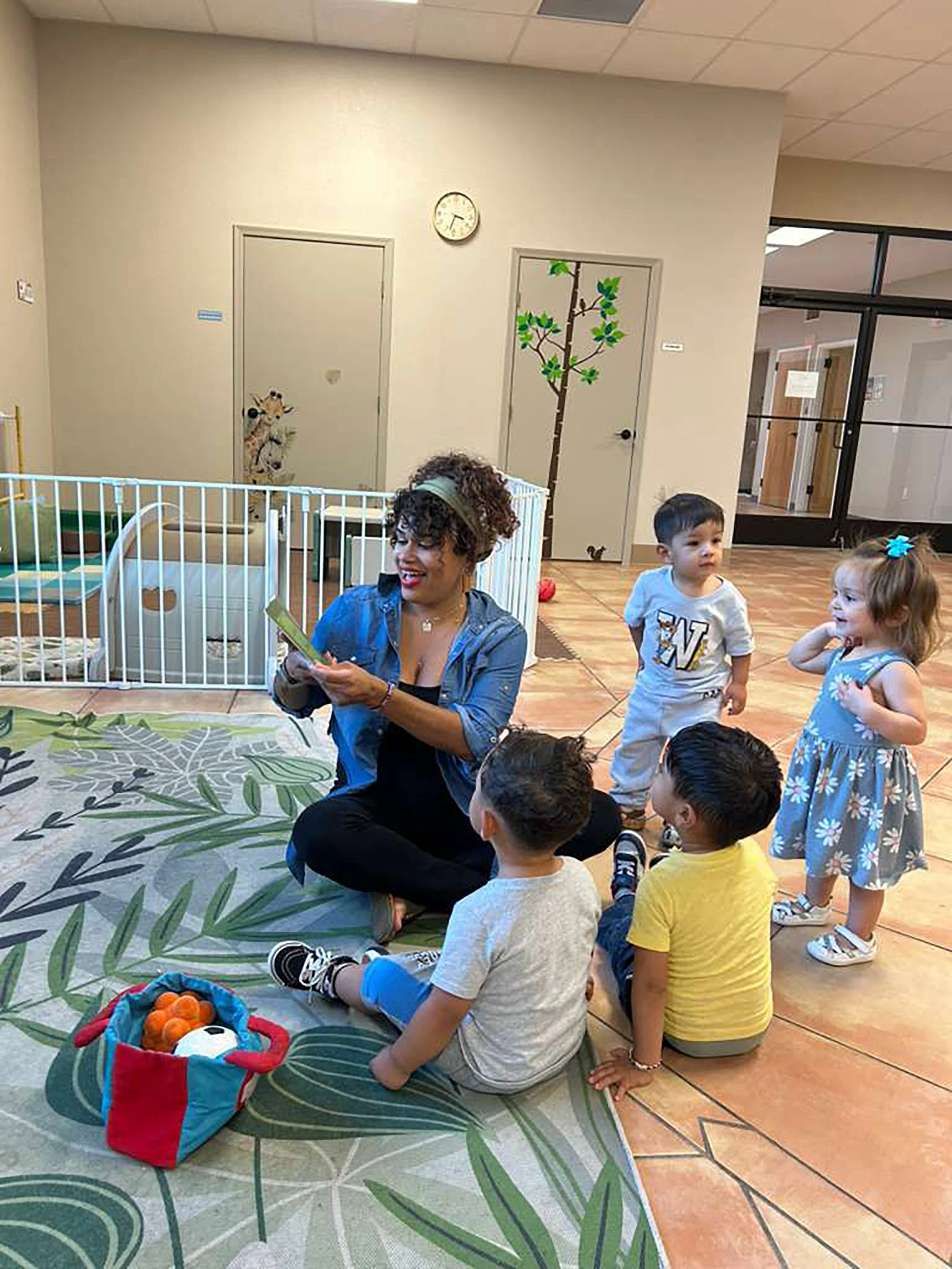 A group of young children gather around a smiling woman reading a bilingual book in a colorful indoor space, decorated with animal and nature-themed wall art, with a cage of fake oranges and a stuffed animal nearby.