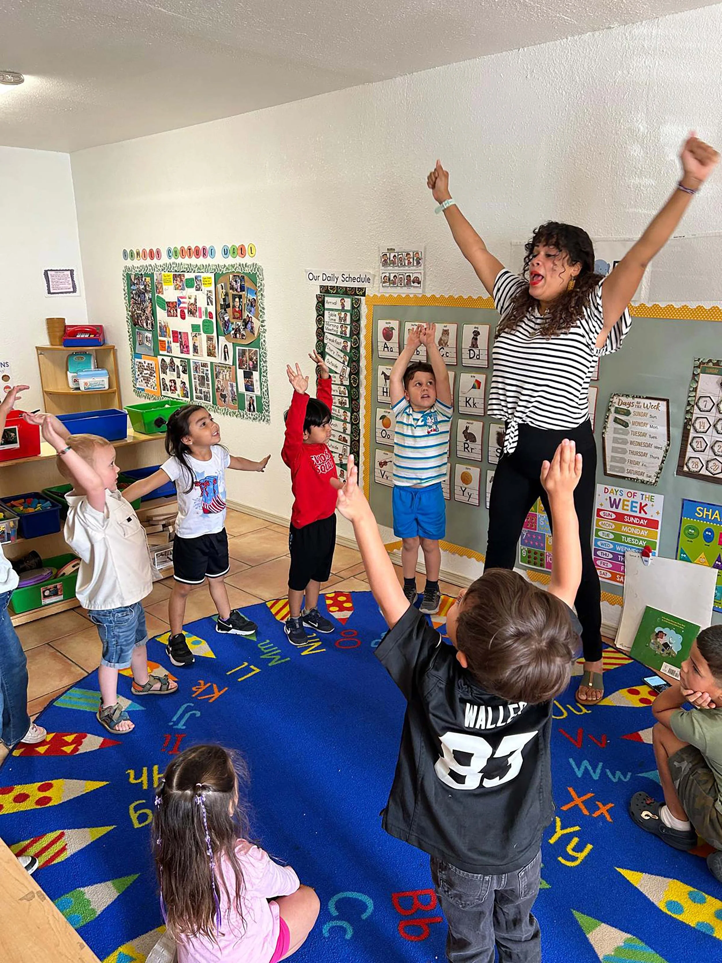 A teacher leading a classroom activity with young children raising their hands, inside a classroom with educational posters and colorful decor.