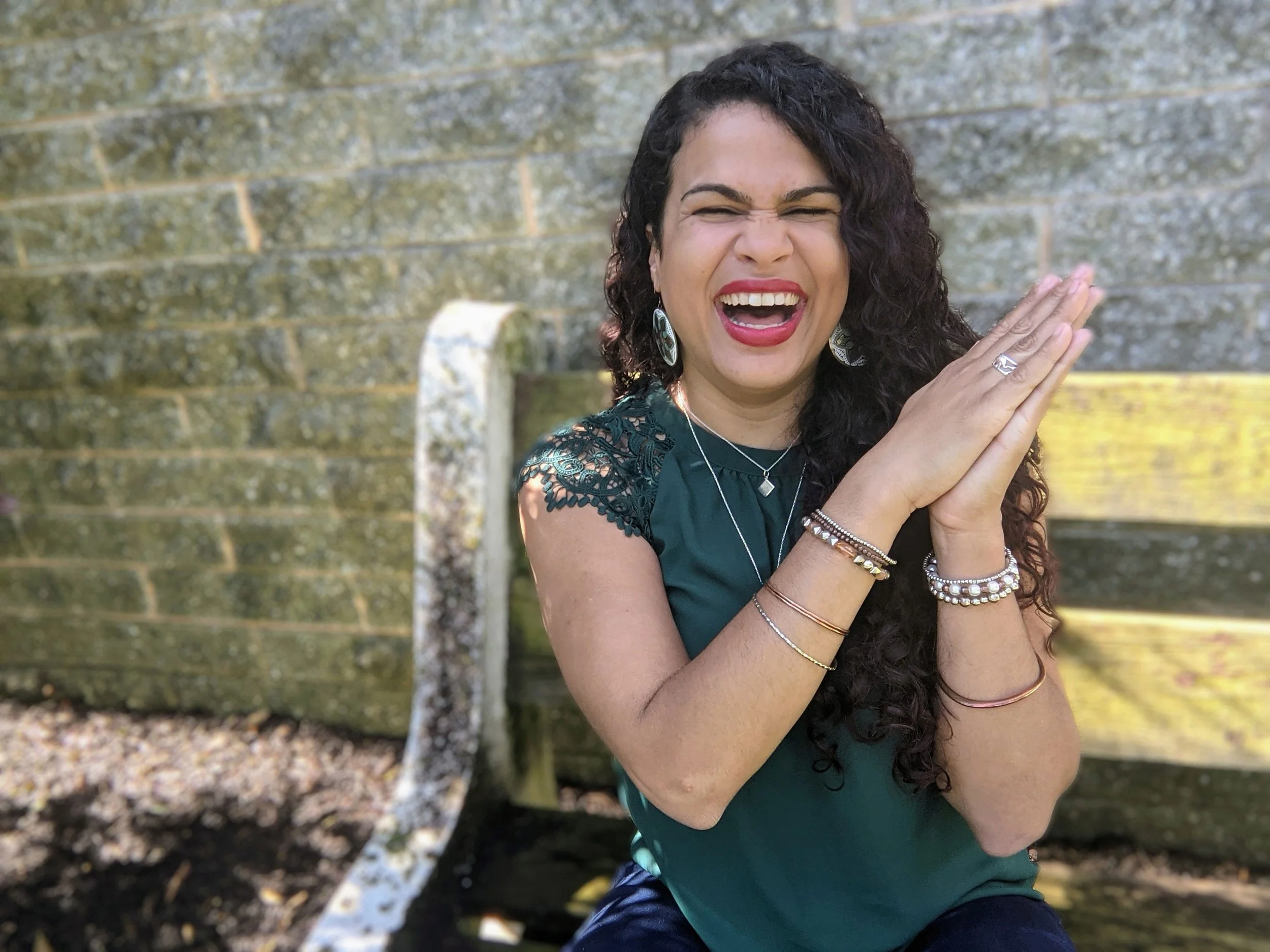 Happy woman with curly dark hair, wearing a dark green dress with lace sleeves and jewelry, laughing with eyes closed, hands pressed together near her face, outdoors with a brick wall and sunlight in the background. This is Jade of ¡Looking Bilingüe!