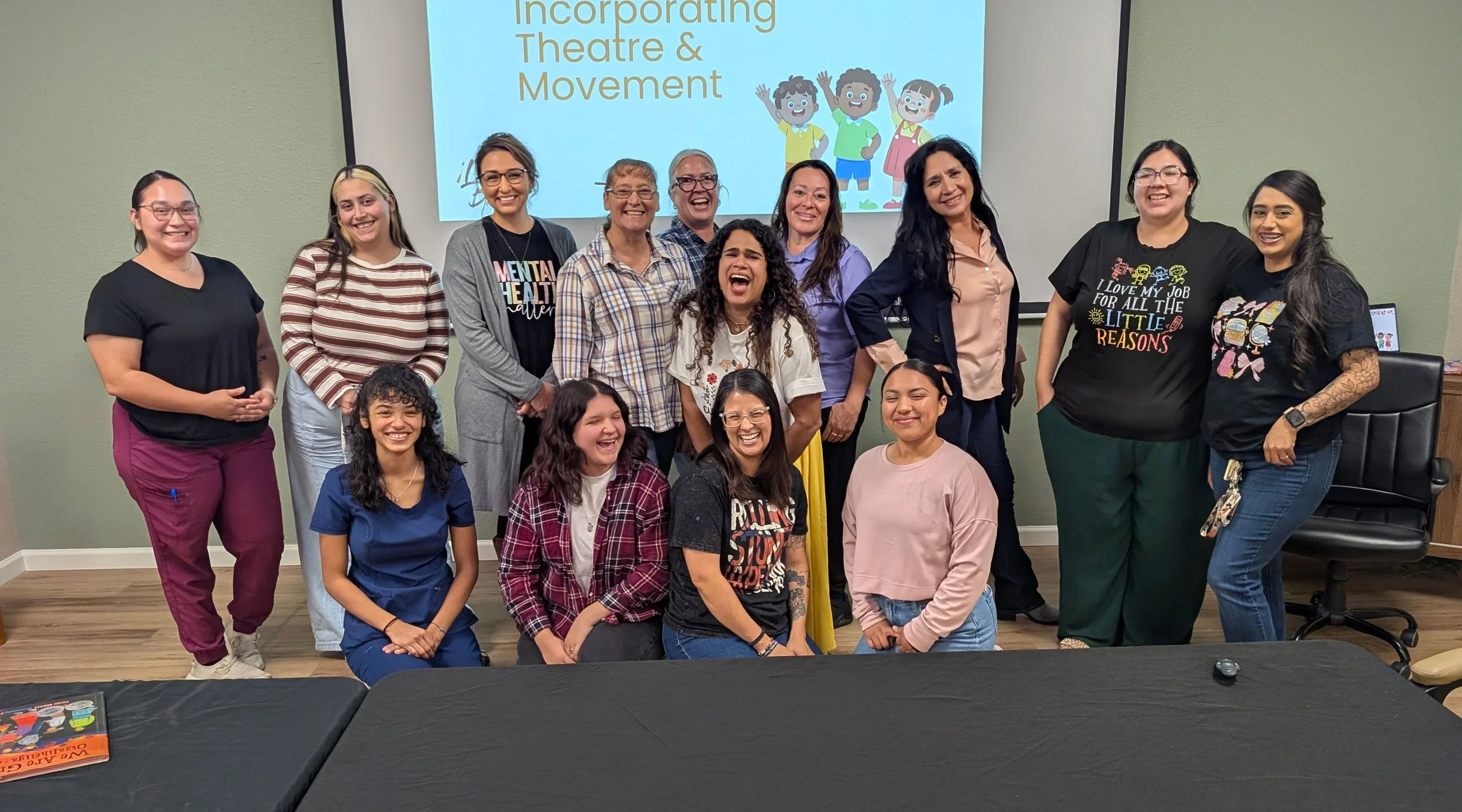 A group of sixteen women and girls standing together in front of a presentation screen that reads 'Incorporating Theatre & Movement' with cartoon children on it. They are smiling and appear happy at a workshop about bilingual learning.
