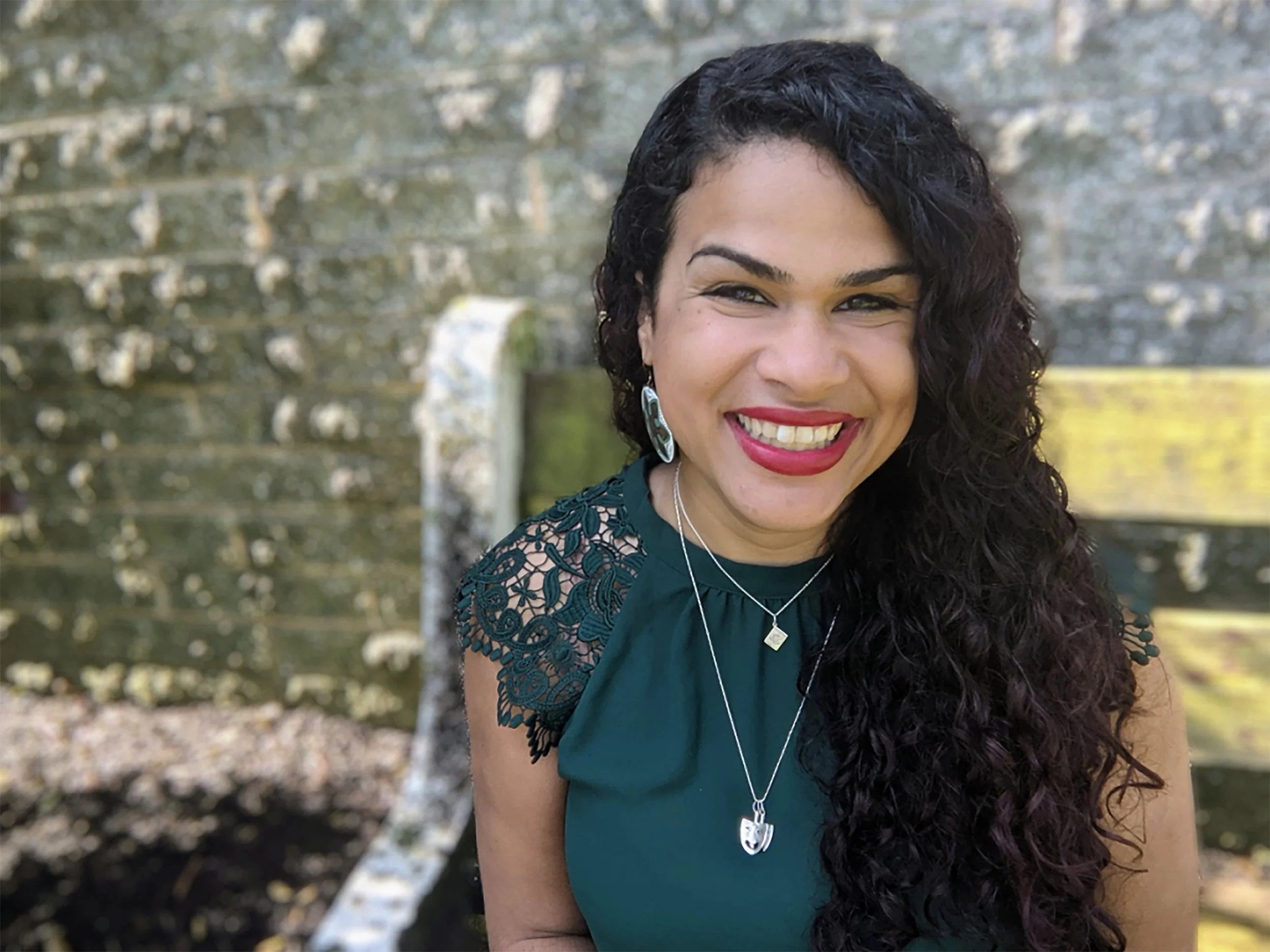 A woman with dark curly hair and red lipstick smiling outdoors in front of trees. This is Jade of ¡Looking Bilingüe!