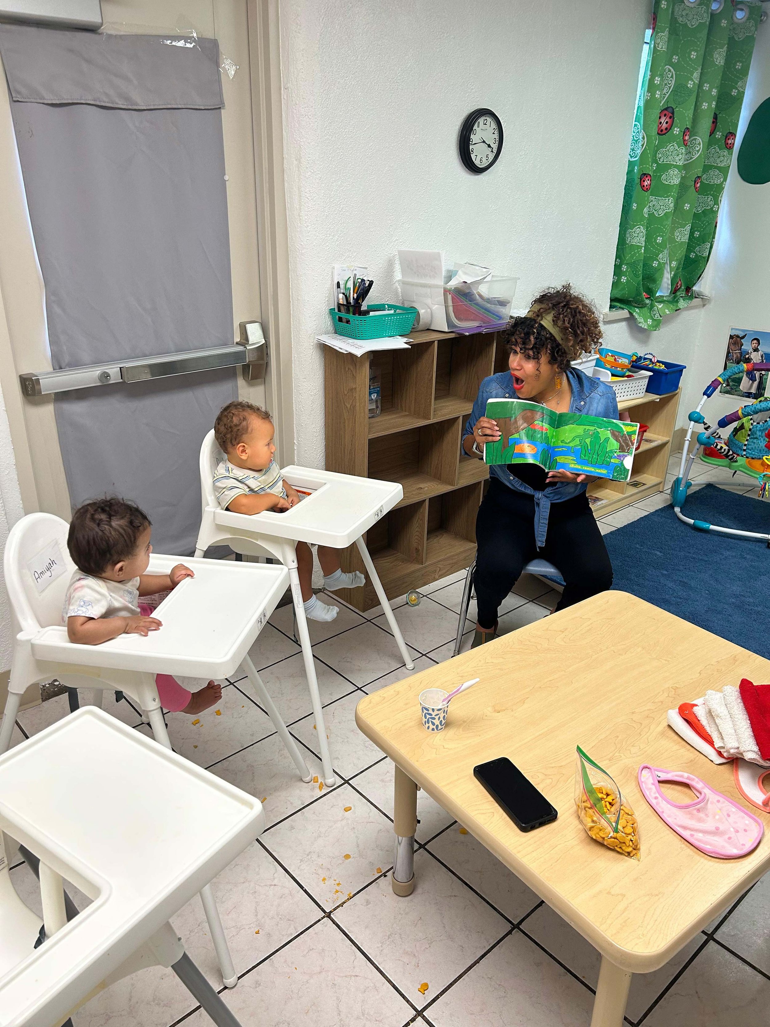 A woman reading a bilingual book to a group of young children in high chairs in a daycare or classroom setting. There is a table with snacks, a phone, and children's items in the foreground, and toys and storage on the shelves in the background.