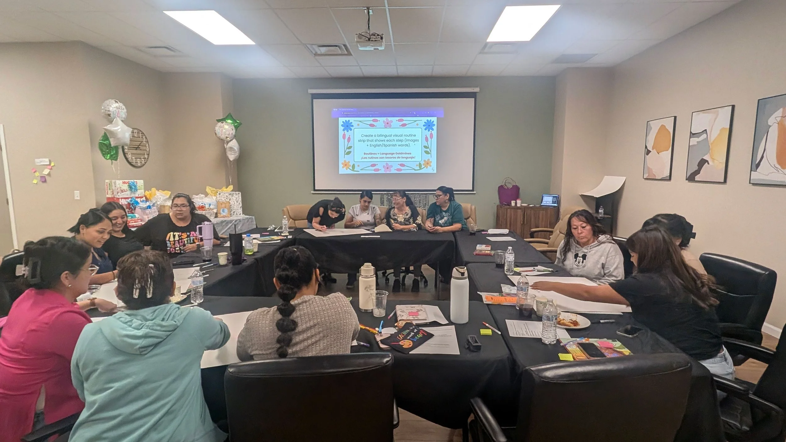 A group of women gathered around a U-shaped conference table in a meeting room, engaged in discussion about bilingual learning with notebooks, papers, and water bottles on the table, with a presentation projected on a screen at the front of the room.