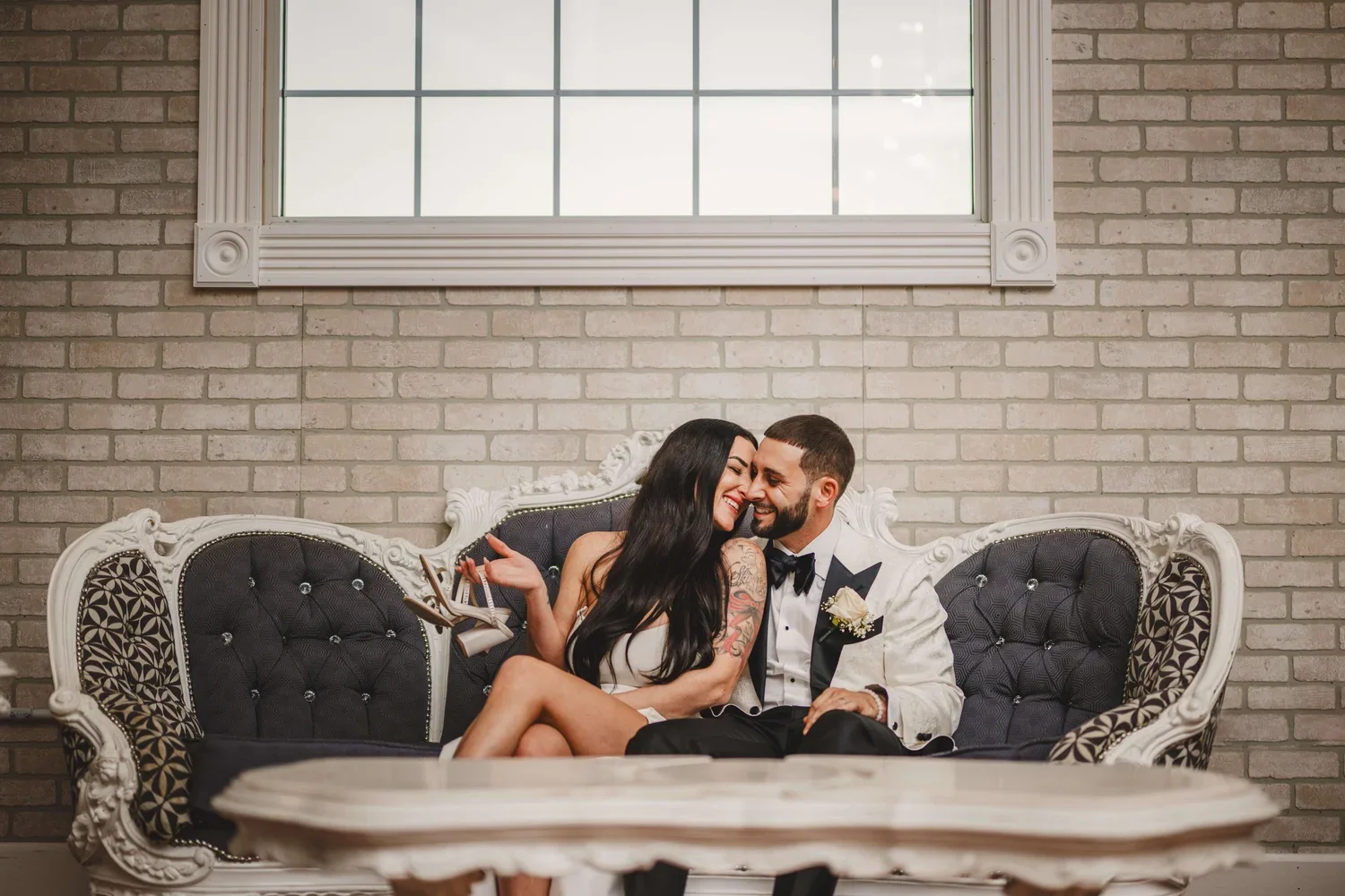 Bride and groom sitting on an antique black couch smiling at each other, with the bride holding her shoes after taking them off.