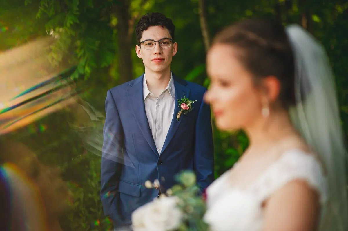 Groom in a blue suit looking directly at the camera in sharp focus, with the bride softly blurred in the foreground during their wedding portraits.