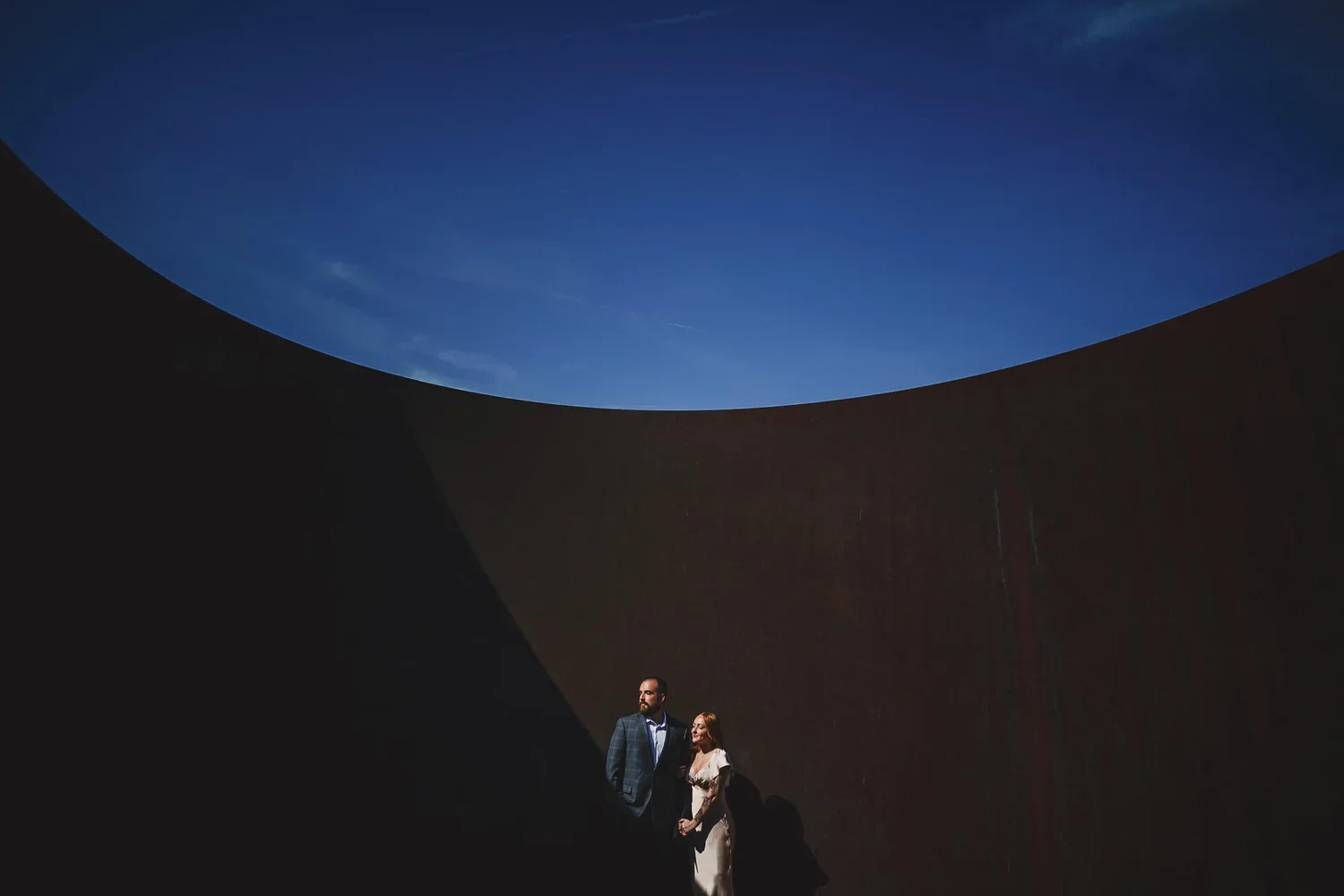 Couple posing during an outdoor engagement photo session with a blue sky and curved dark wall in the background.