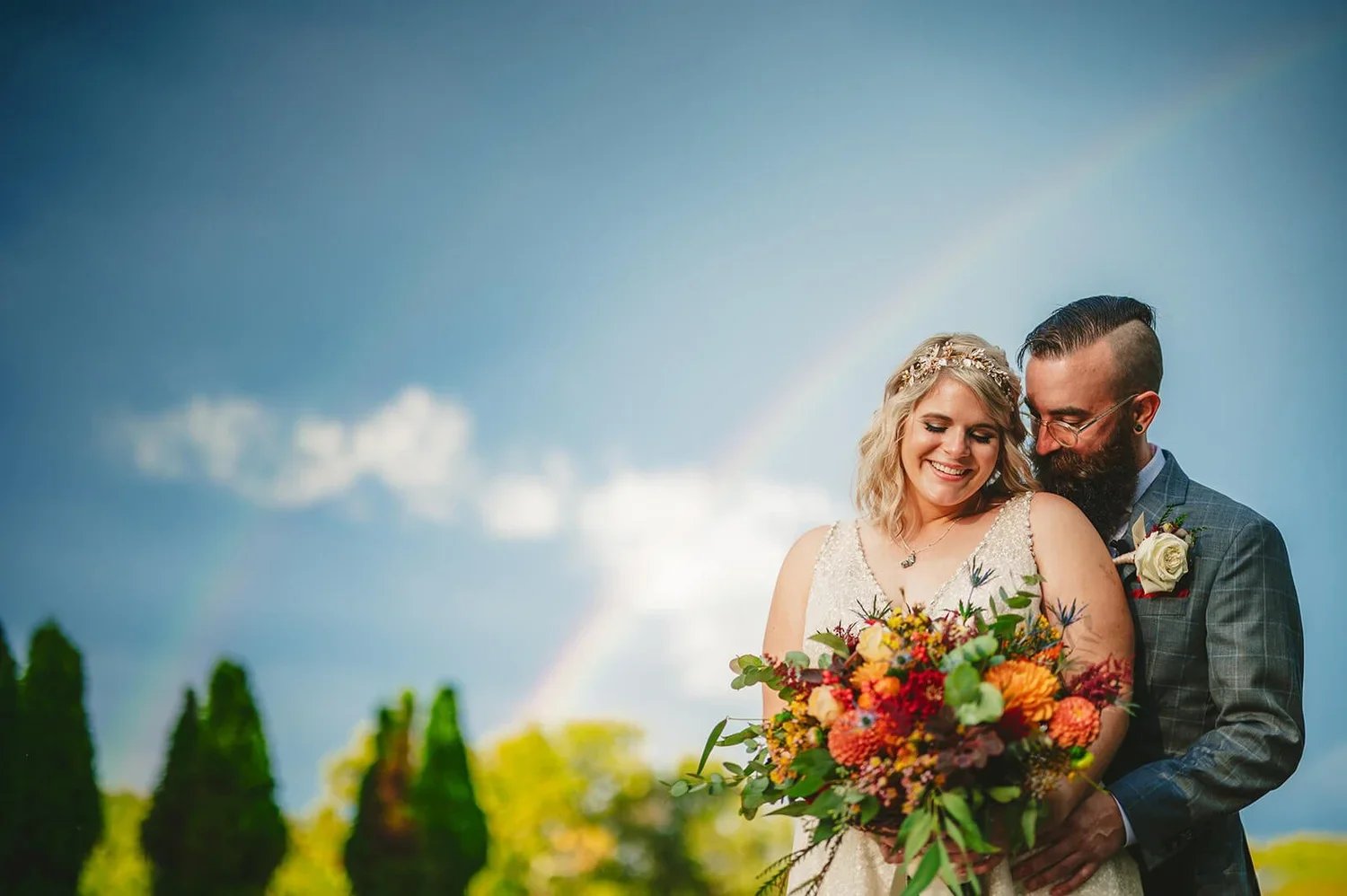 Colorful outdoor wedding portrait of a bride and groom embracing and smiling, with the bride holding a vibrant bouquet of wedding flowers.