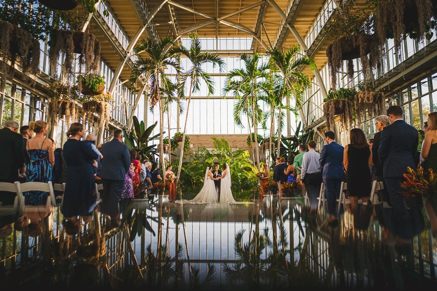 two brides at the end of the aisle, facing each other in a greenhouse wedding venue with tall greenery surrounding them