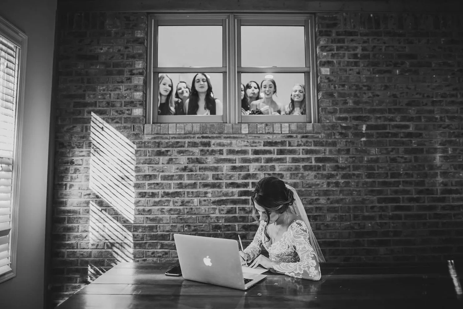 Black and white wedding photo of a bride writing something down with her laptop nearby while guests watch from above and smile.