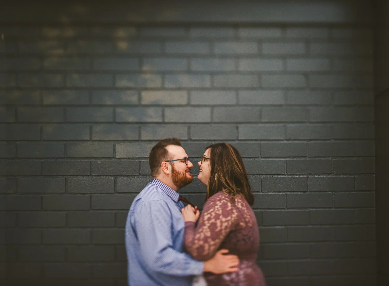Couple facing each other and smiling in front of a dark brick wall during their engagement photoshoot.