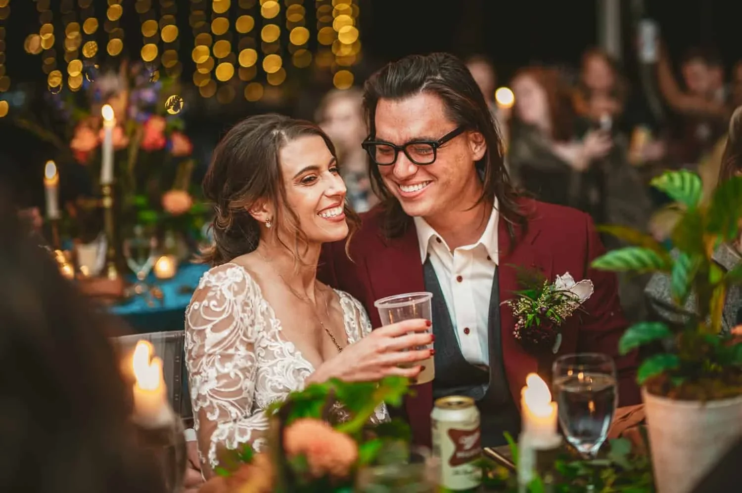Newlyweds sharing drinks and smiling during their wedding reception, with the groom dressed in a burgundy suit.