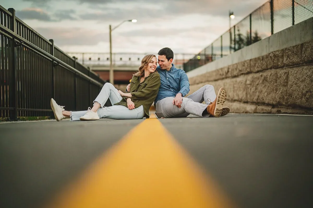 Couple sitting side by side outdoors, smiling and looking at each other with a yellow road line centered between them.
