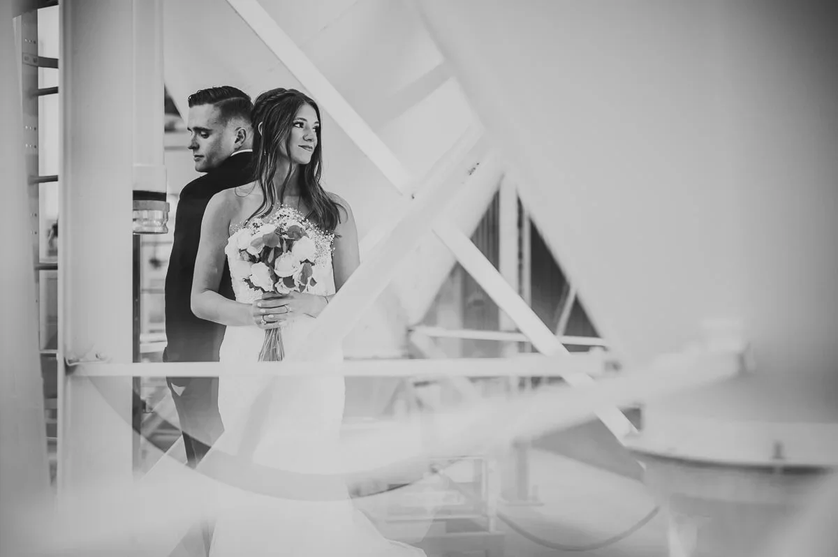Black and white wedding portrait of a bride and groom standing back to back in their wedding attire, with the bride holding a bouquet.