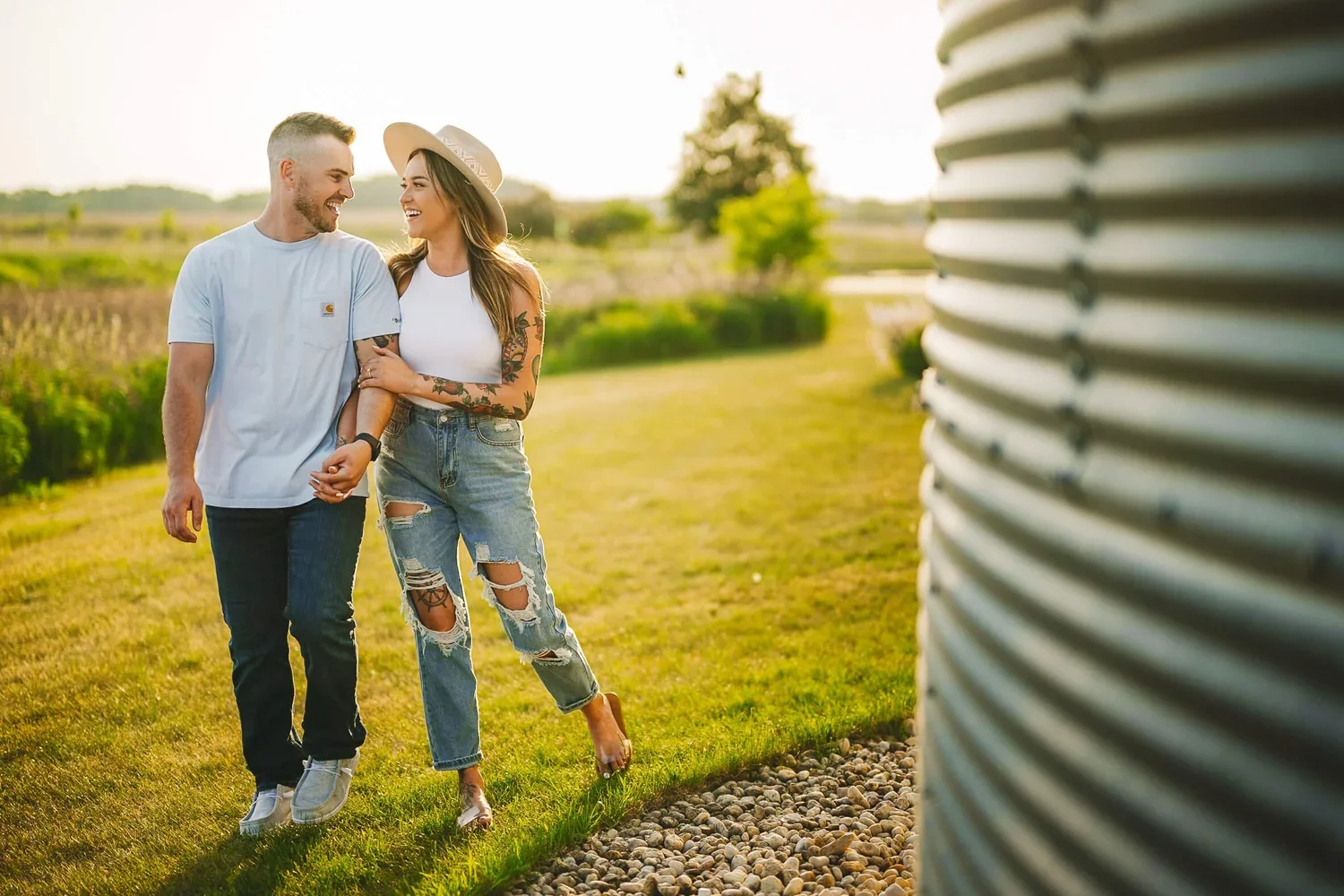 Couple smiling and looking at each other during an outdoor engagement photoshoot, both wearing jeans with one partner wearing a hat.