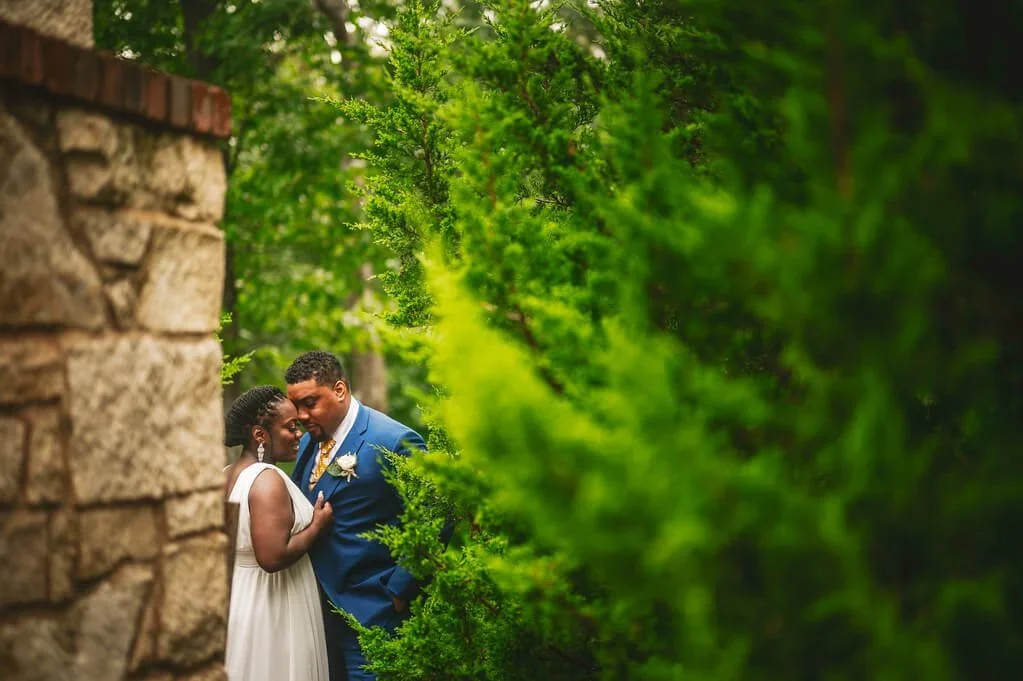 Bride and groom touching foreheads while facing each other outdoors, surrounded by lush greenery.