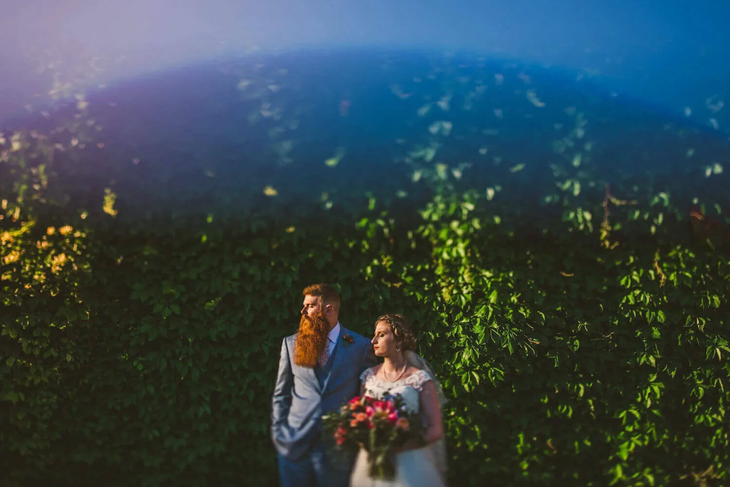 Bride and groom looking to the left in their wedding attire, with the bride holding a bright floral bouquet against a lush green plant-filled background.