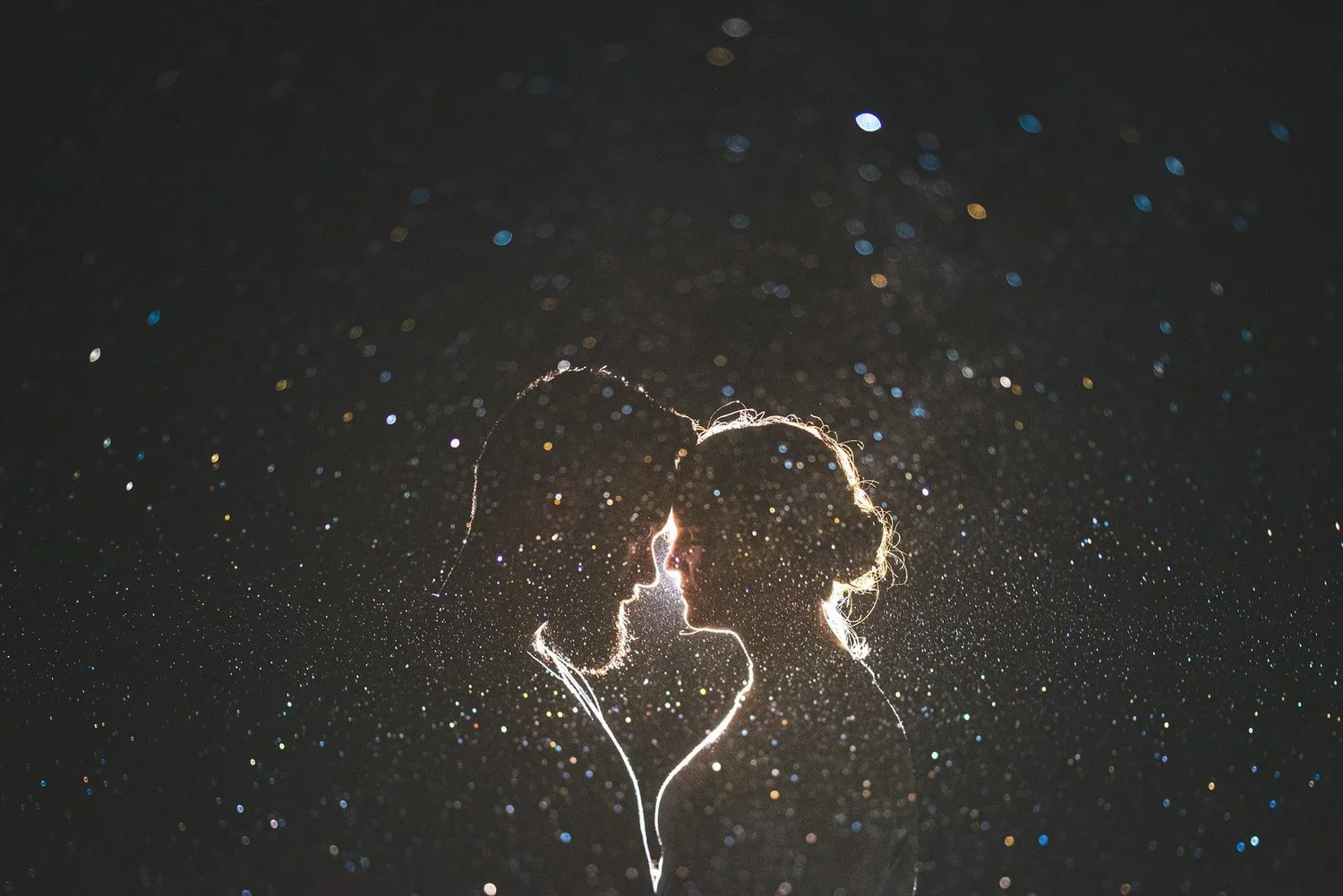Silhouette of a couple facing each other with glowing light outlining them, surrounded by sparkling lights against a dark background.