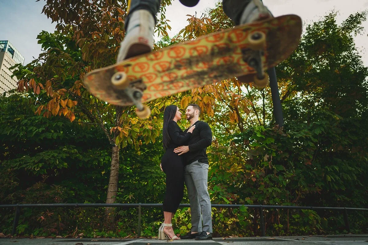 Couple embracing and looking at each other in focus while a skateboarder jumps in the blurred foreground.