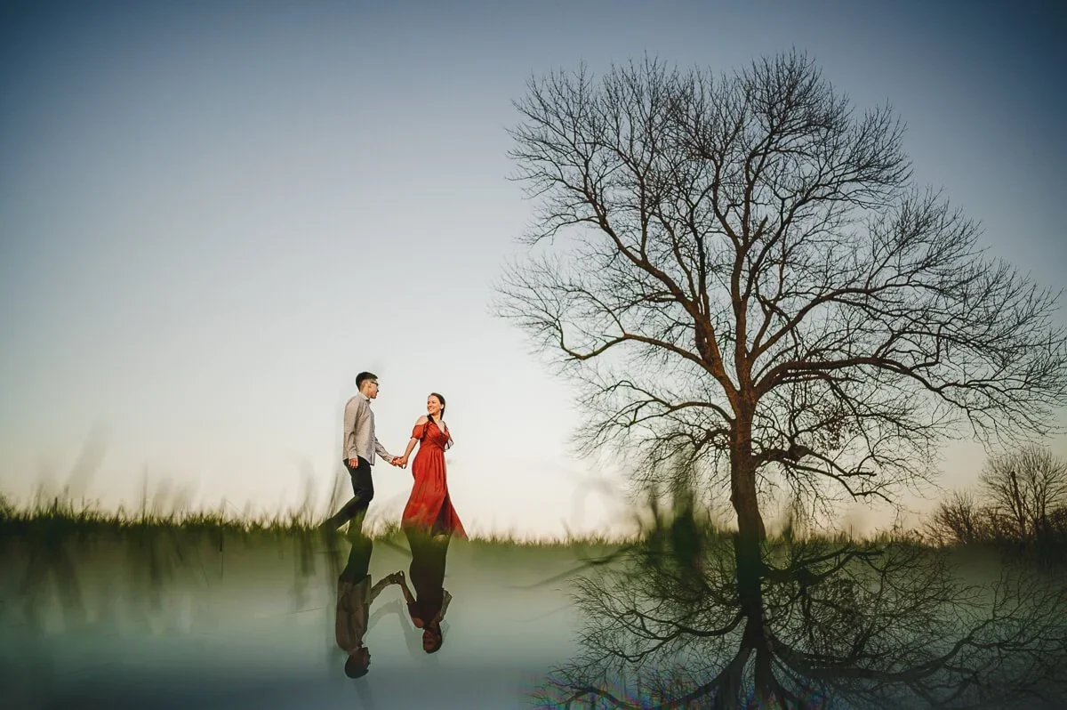 Couple walking hand in hand with one partner wearing a red dress, a tree on the right side, and a mirrored reflection creating the bottom half of the image.