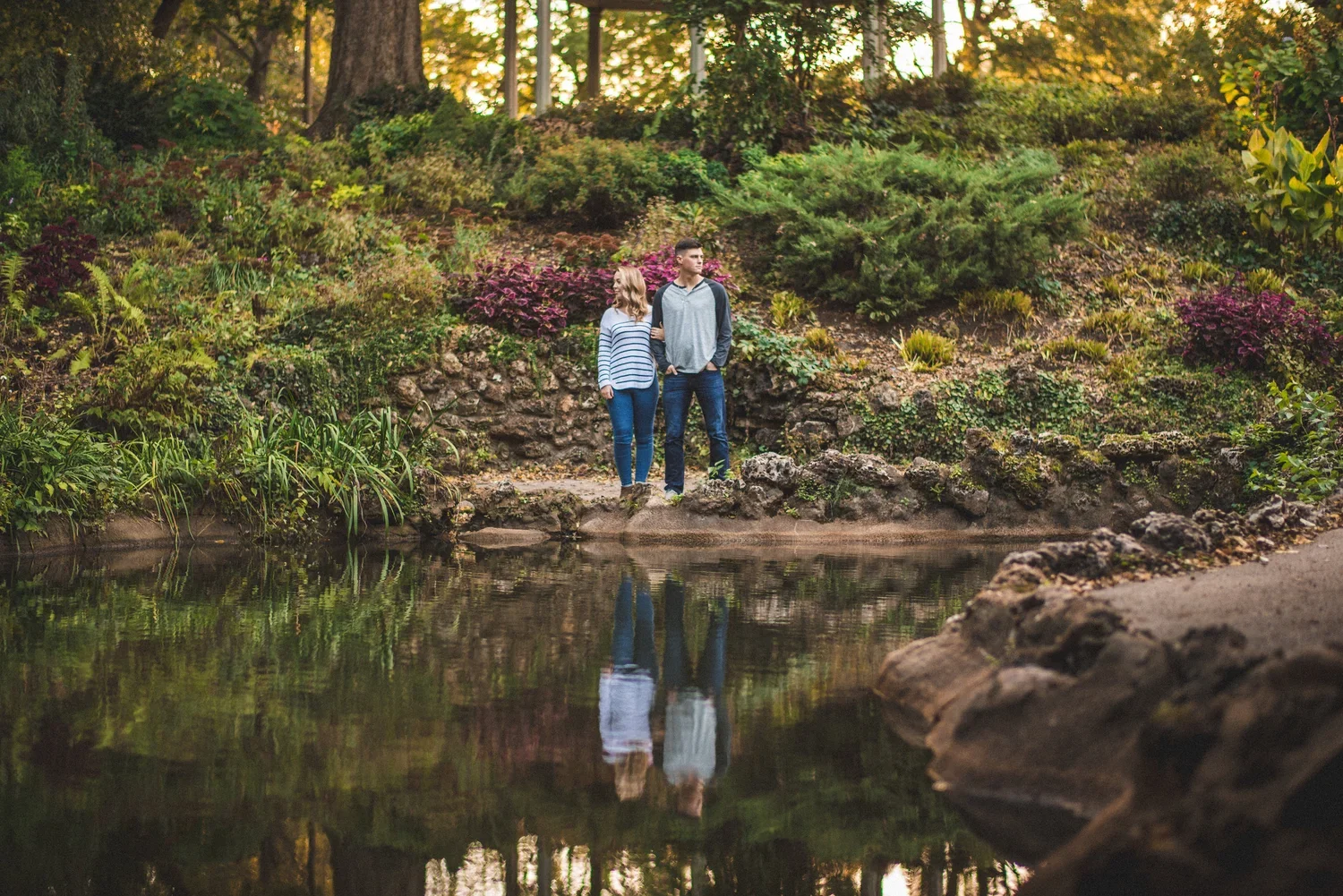 Engaged couple standing outdoors surrounded by greenery and water, looking in opposite directions while linking arms.