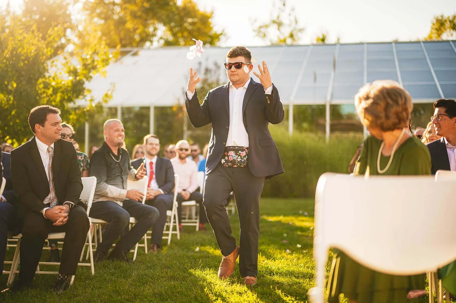 Groomsman walking down the aisle on the wedding day while tossing items in the air, wearing a fanny pack as guests laugh.
