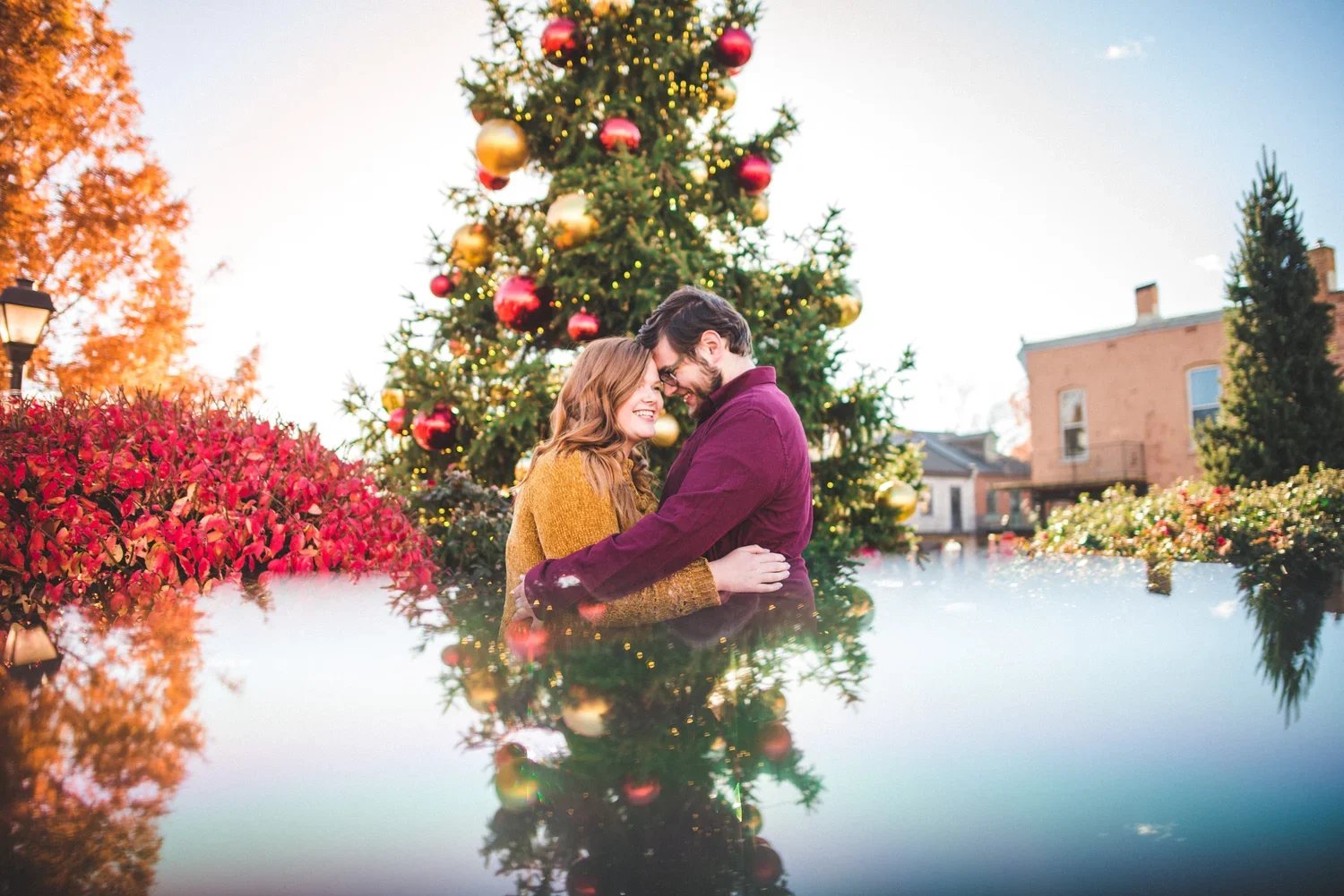 Couple posing in front of a decorated Christmas tree with ornaments, with the bottom half of the image showing a reflection of the tree.