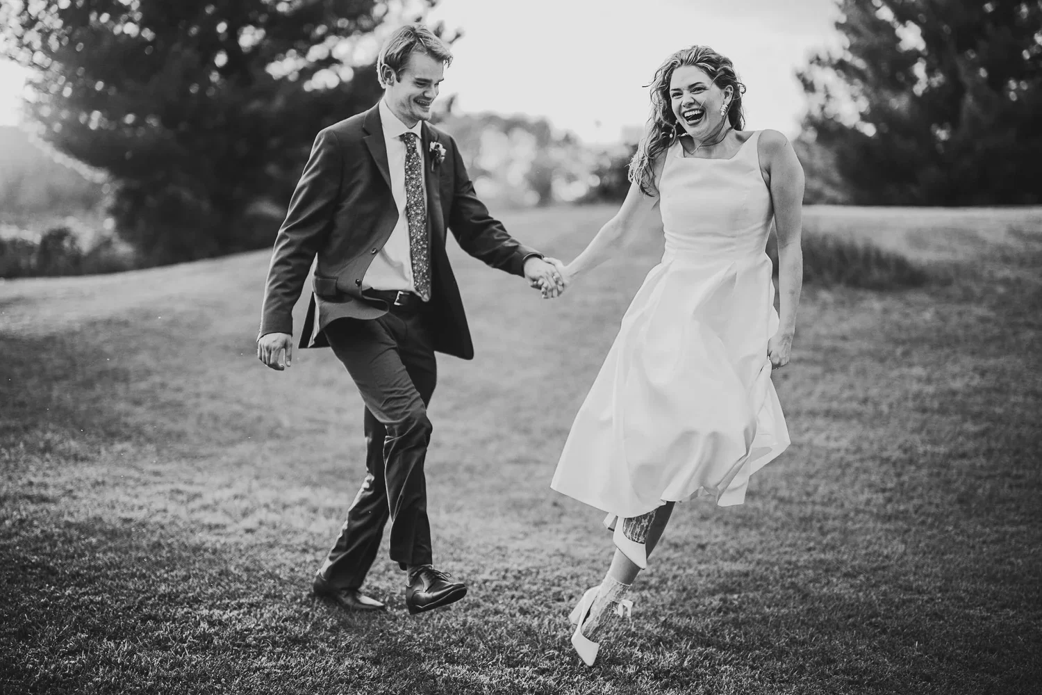 Black and white wedding portrait of a couple holding hands, smiling, and skipping together outdoors in their wedding attire.