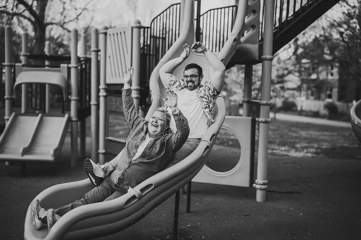 Black and white image of a couple sliding down a playground slide with their arms raised and smiling.