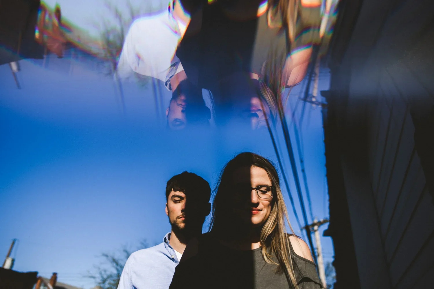 Double exposure engagement portrait of a couple with dramatic shadows across their faces beneath a bright blue sky.