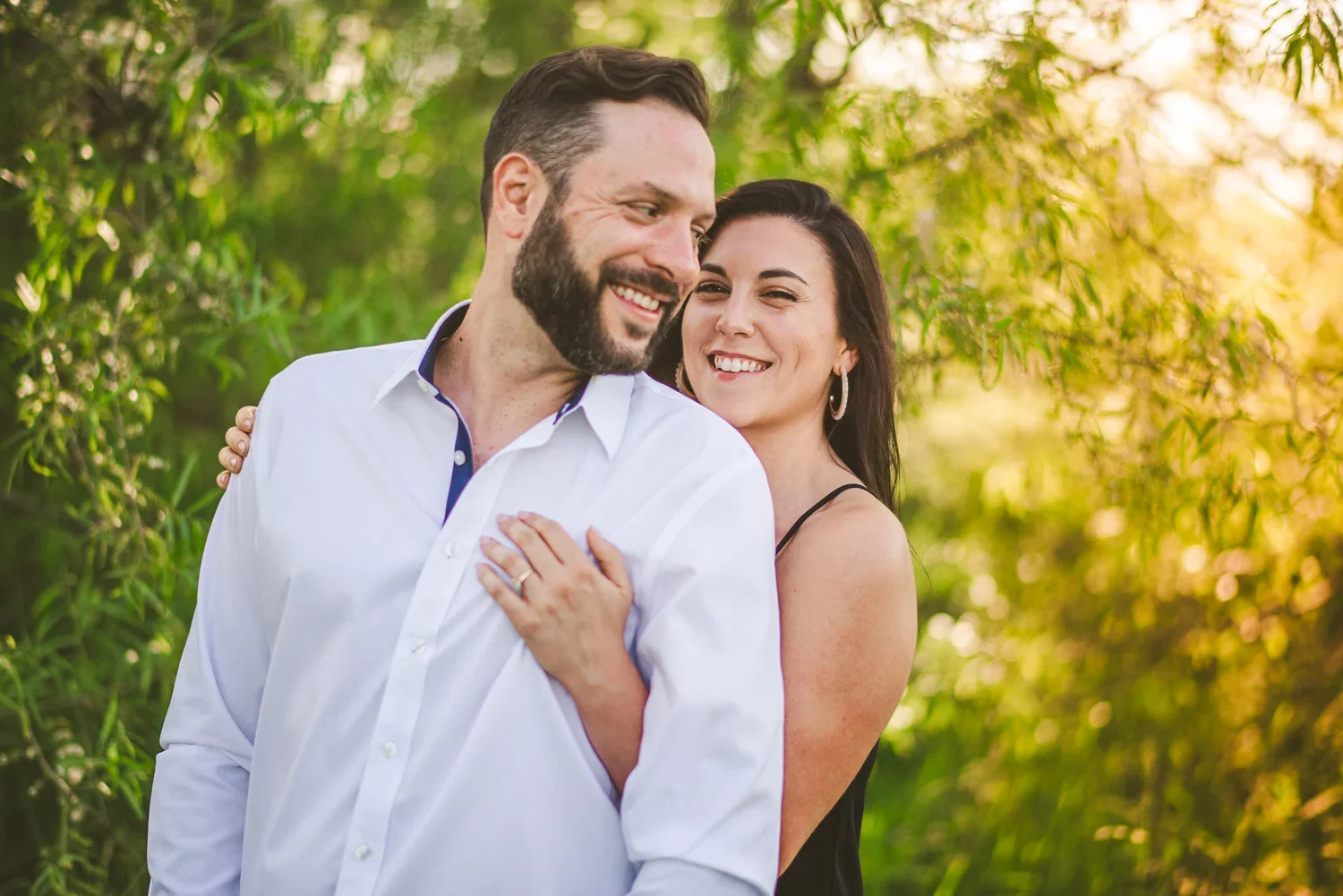 Engagement portrait of a couple hugging and smiling together surrounded by trees and foliage.