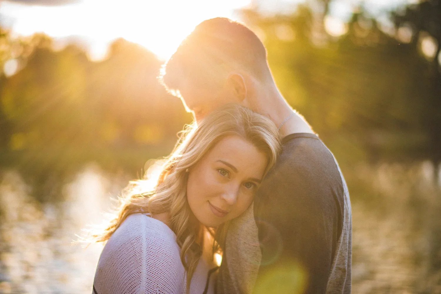 Engagement portrait during golden hour with the woman looking directly at the camera and her partner softly blurred in the background.