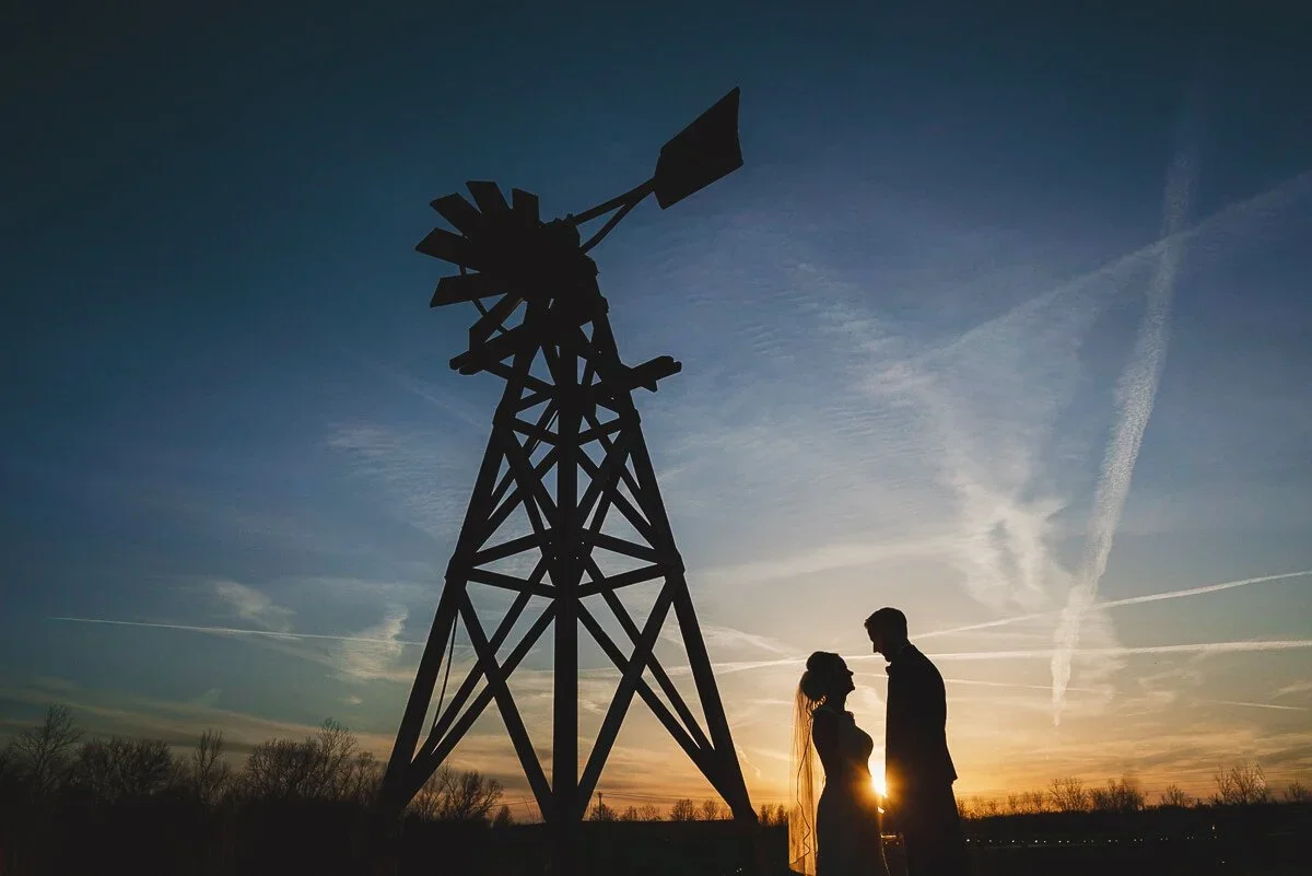 Silhouette of a bride and groom standing beside a windmill at sunset with a blue evening sky.