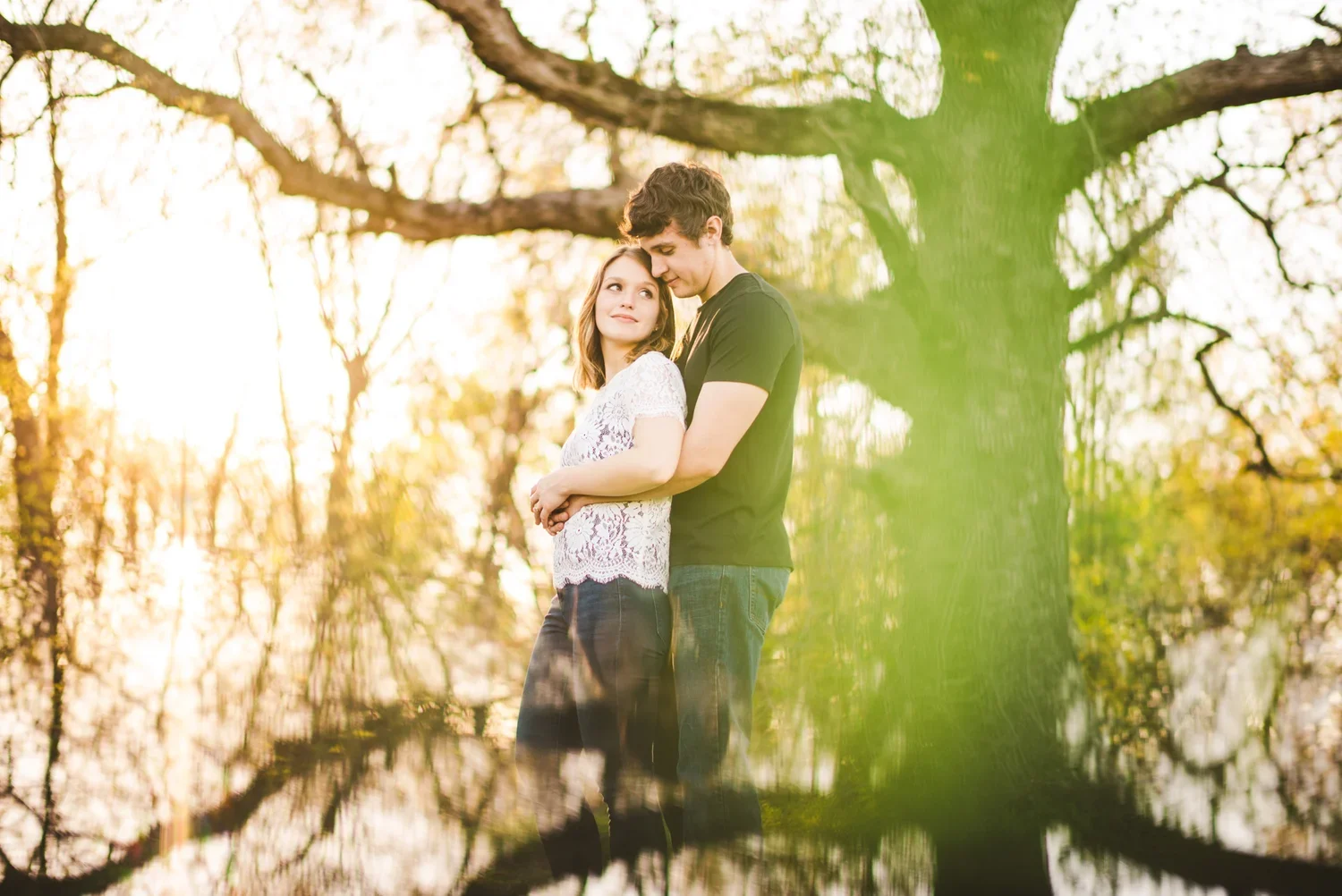 Couple posing outdoors during an engagement photoshoot surrounded by trees, with a mirrored reflection creating the bottom half of the image.
