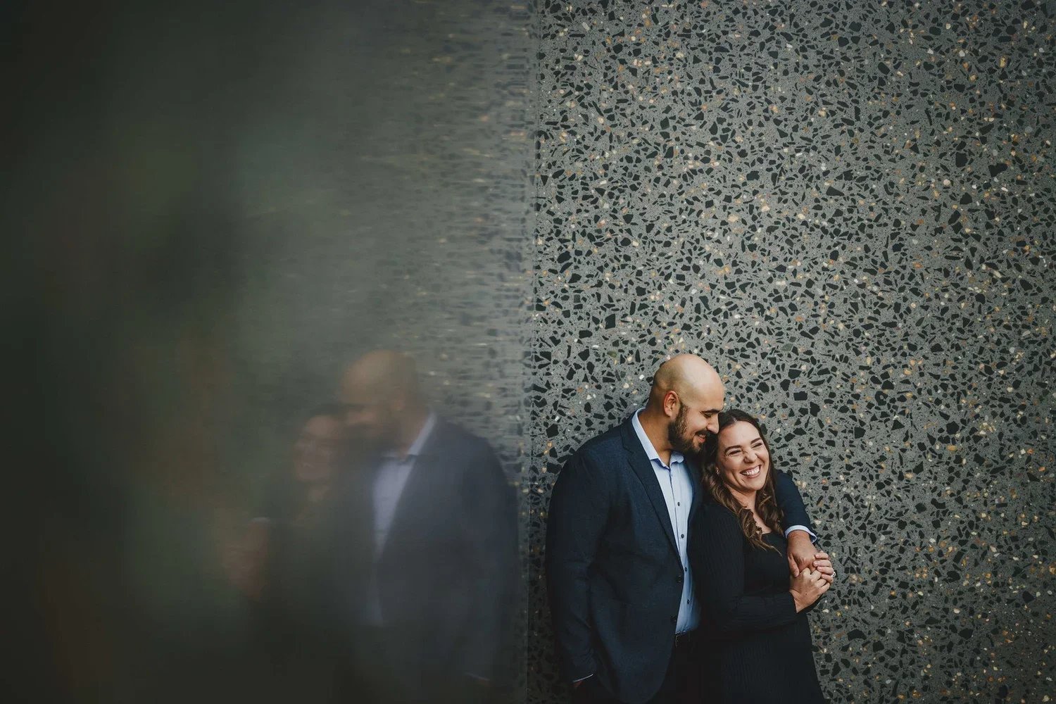 Engaged couple smiling with their arms around each other while looking away from the camera against a dark background.