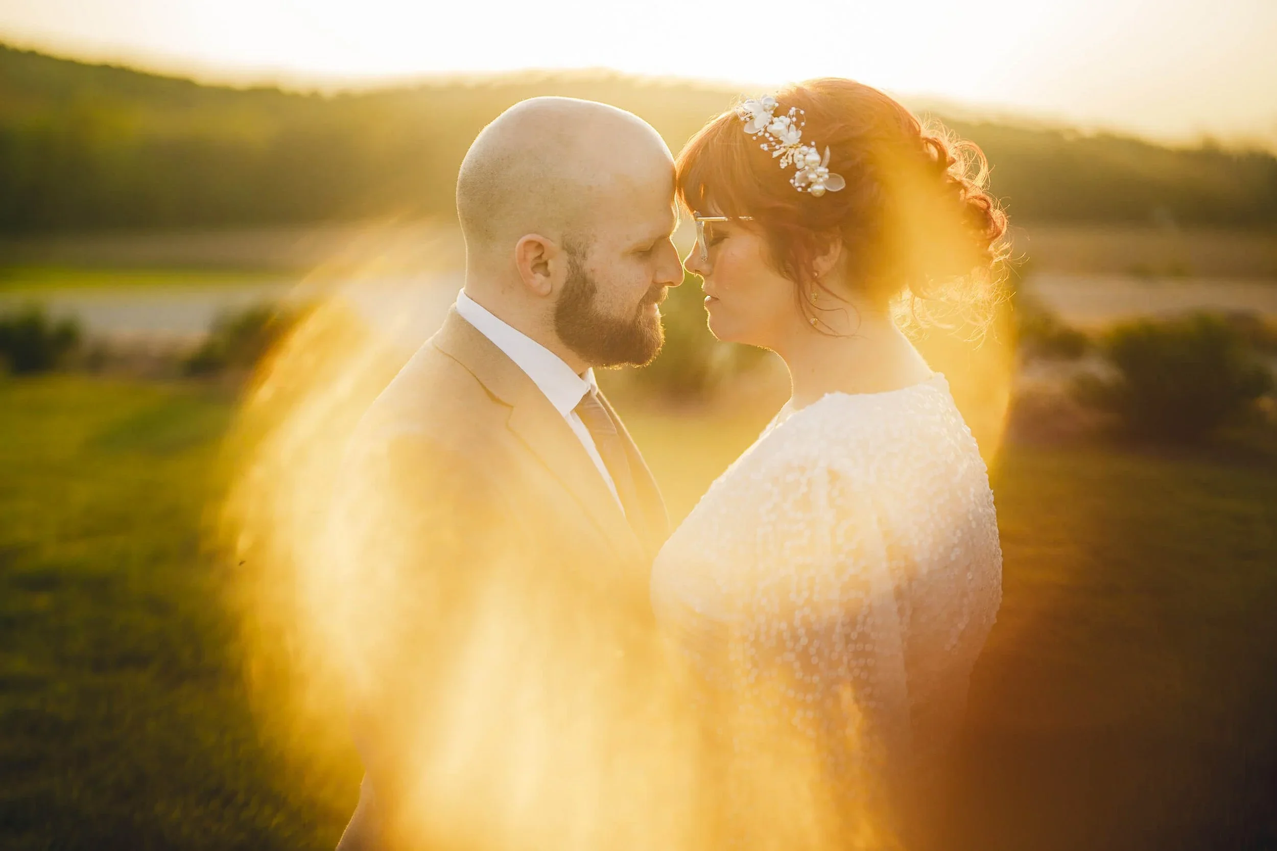 Bride and groom facing each other in wedding attire outdoors during golden hour with a warm sun flare between them.