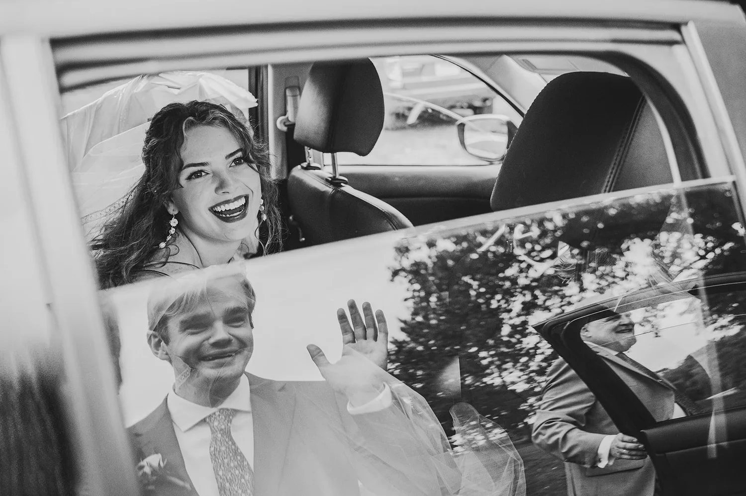 Black and white wedding photo of a bride and groom, with the bride in a car with the window down and the groom waving in the reflection.